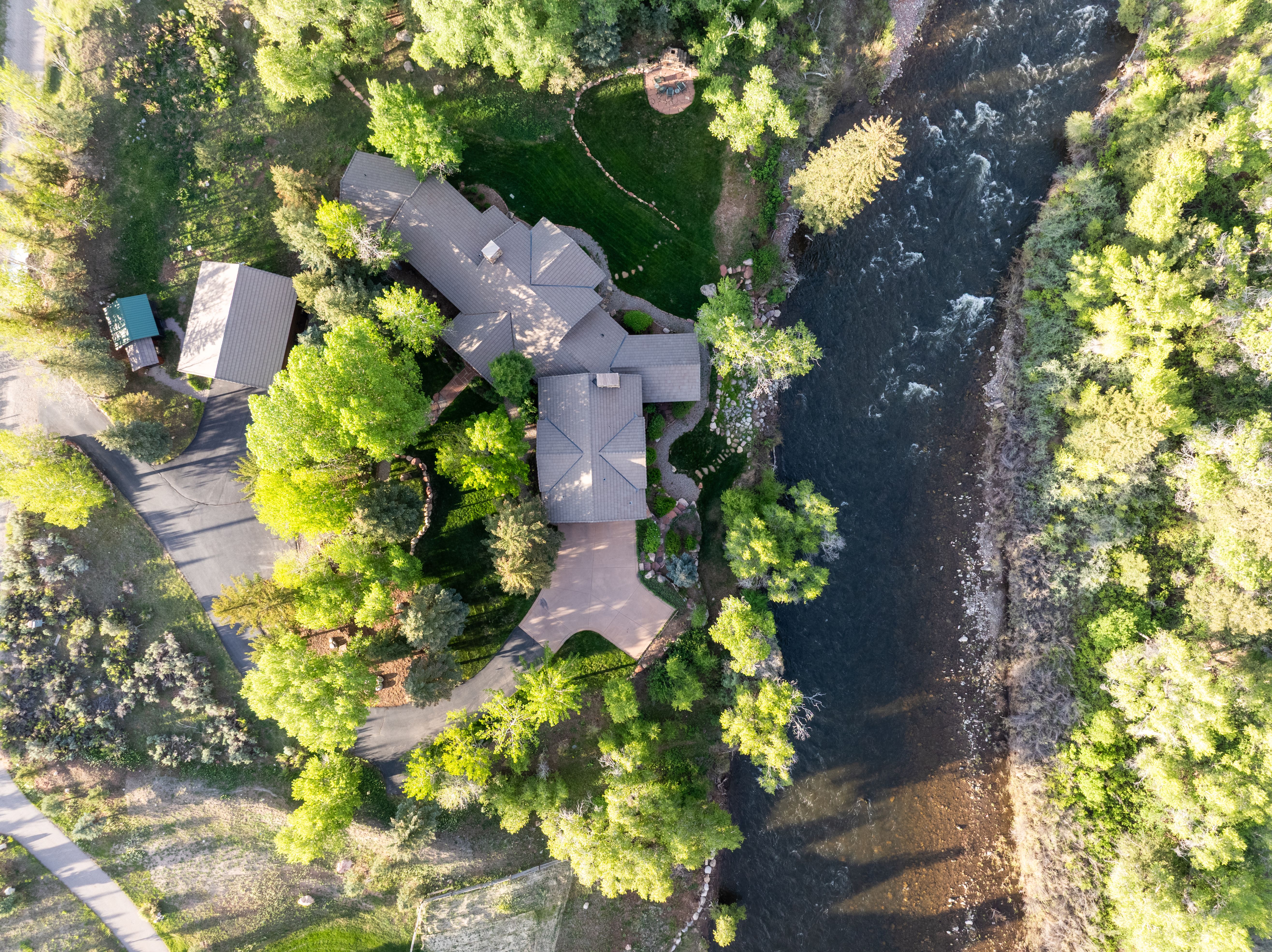 This high-angle aerial view captures a secluded residential property nestled along the banks of a flowing river. The home features a multi-gabled roofline, a paved driveway, and a detached garage, all surrounded by lush green trees and manicured landscaping. The perspective emphasizes the serene, natural setting and the expansive outdoor space, highlighting the property's unique waterfront location.