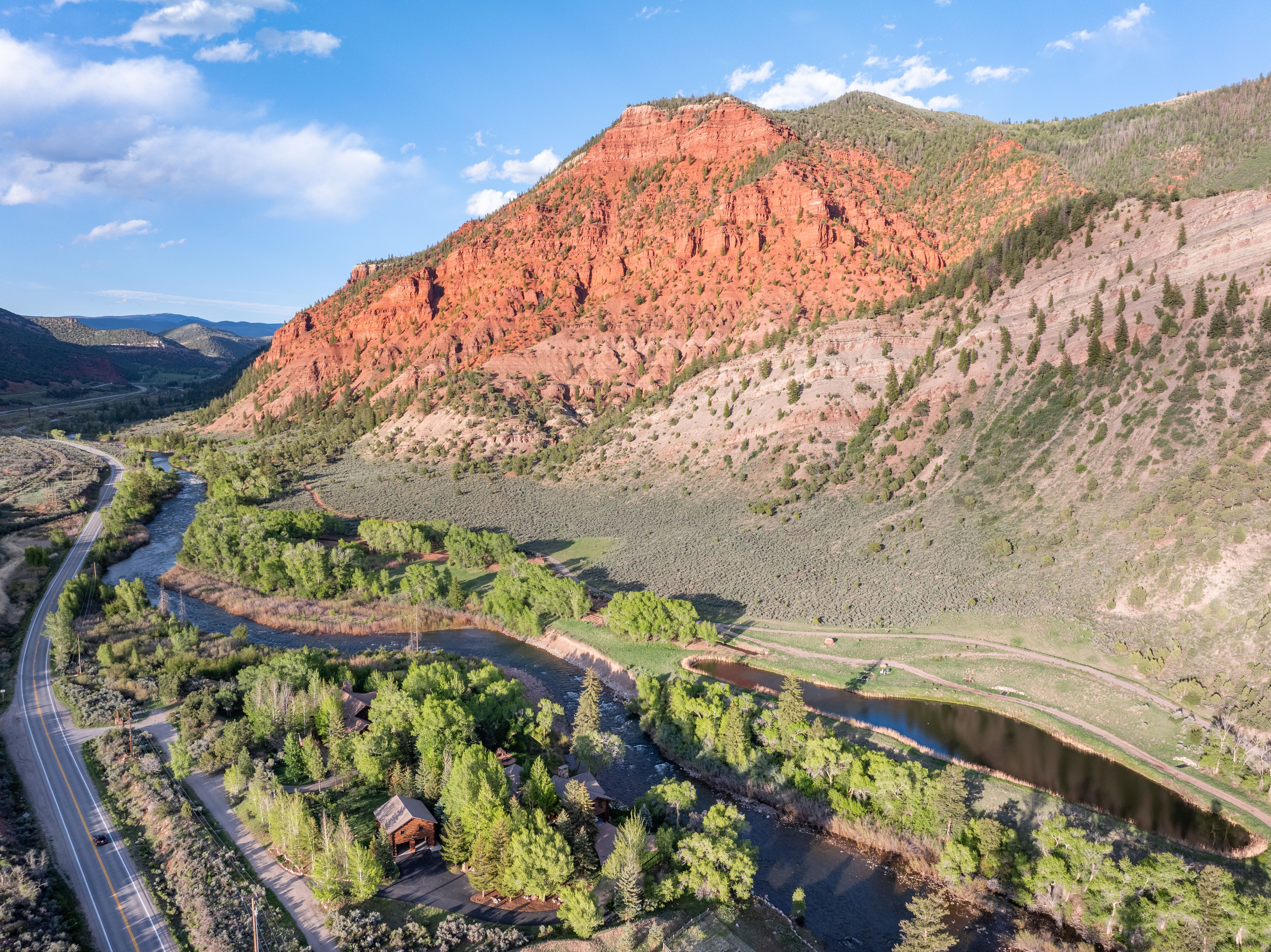 This stunning aerial view captures a rustic cabin nestled within a lush, tree-lined riverbank at the base of a dramatic, towering red rock mountain. The landscape features a winding river, a small pond, and a scenic road running parallel to the property, emphasizing its secluded and picturesque location. The perspective provides a grand, cinematic sense of scale, highlighting the property's integration into the rugged natural beauty of the surrounding terrain.