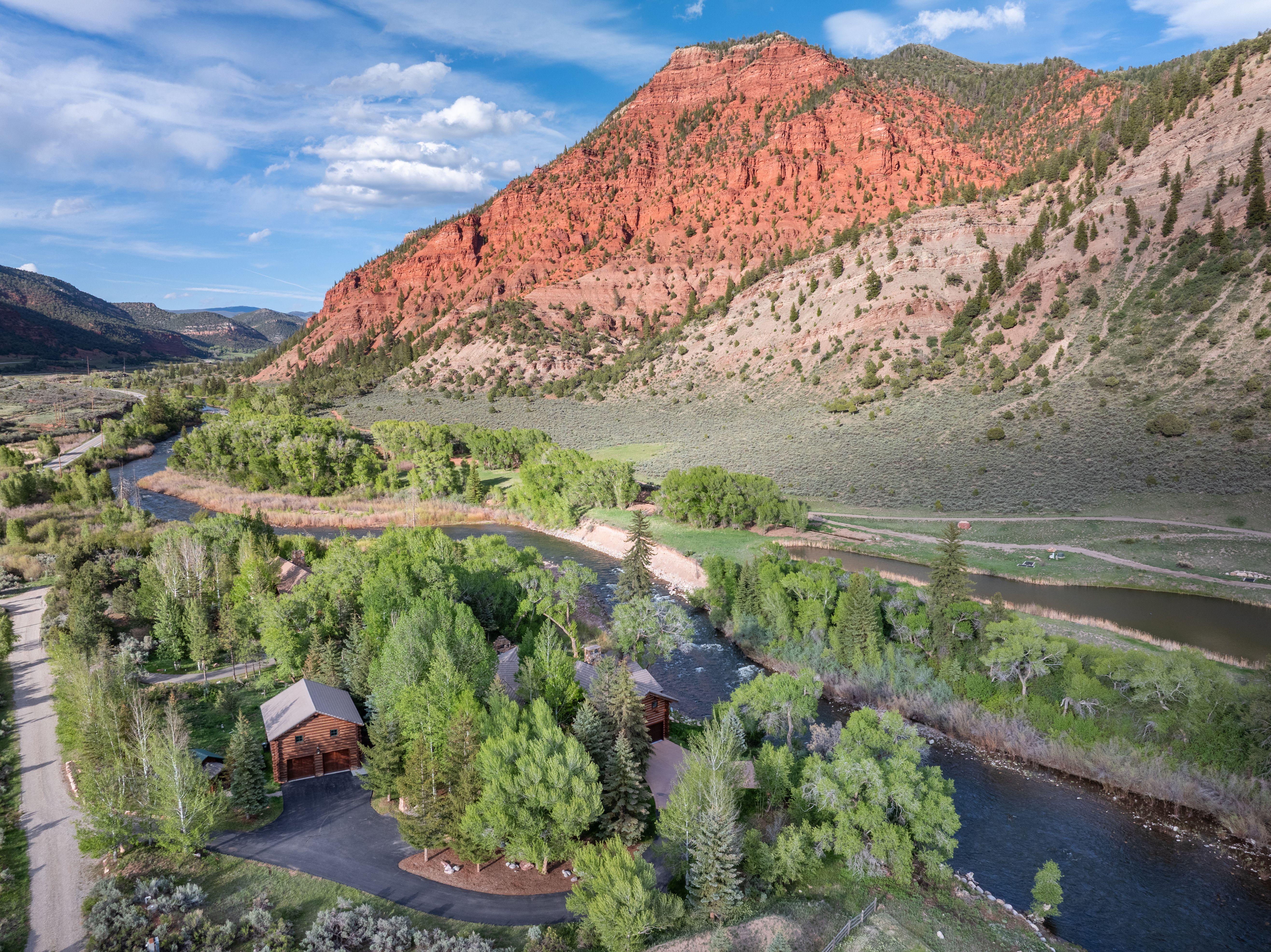 This stunning aerial perspective captures a rustic log cabin nestled along a winding river, set against the dramatic backdrop of a towering, vibrant red rock mountain. The property features a paved driveway, lush green trees, and expansive natural surroundings, emphasizing a serene and secluded mountain retreat. The composition highlights the harmonious blend of architectural craftsmanship and the rugged beauty of the surrounding landscape.