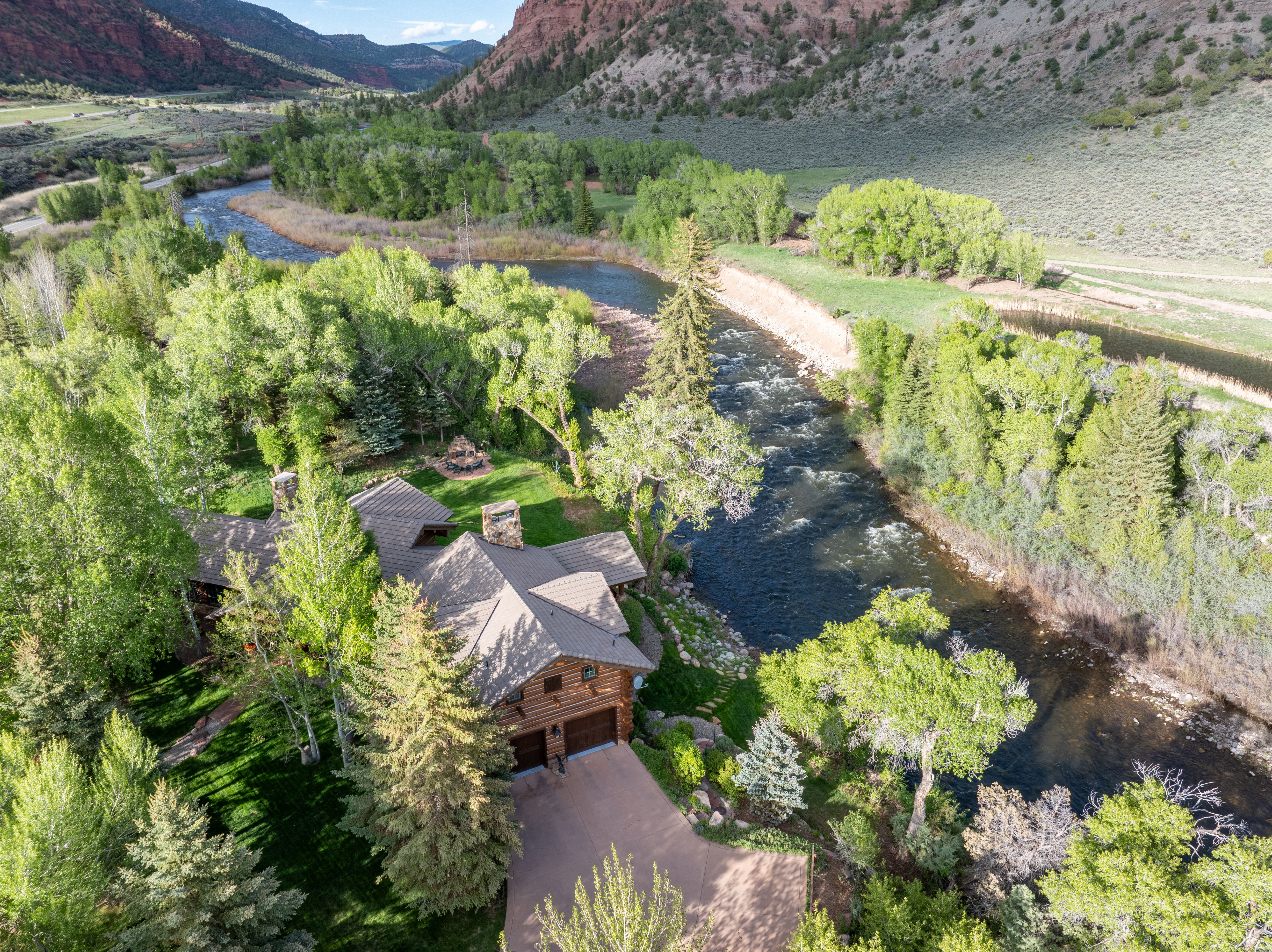 This high-angle aerial shot captures a rustic log cabin nestled along the banks of a winding river in a scenic, mountainous landscape. The property features a well-manicured lawn, a stone patio area, and a paved driveway leading to a two-car garage, all surrounded by lush green trees and dramatic red rock cliffs. The perspective emphasizes the tranquil, secluded nature of the home and its integration into the natural environment.