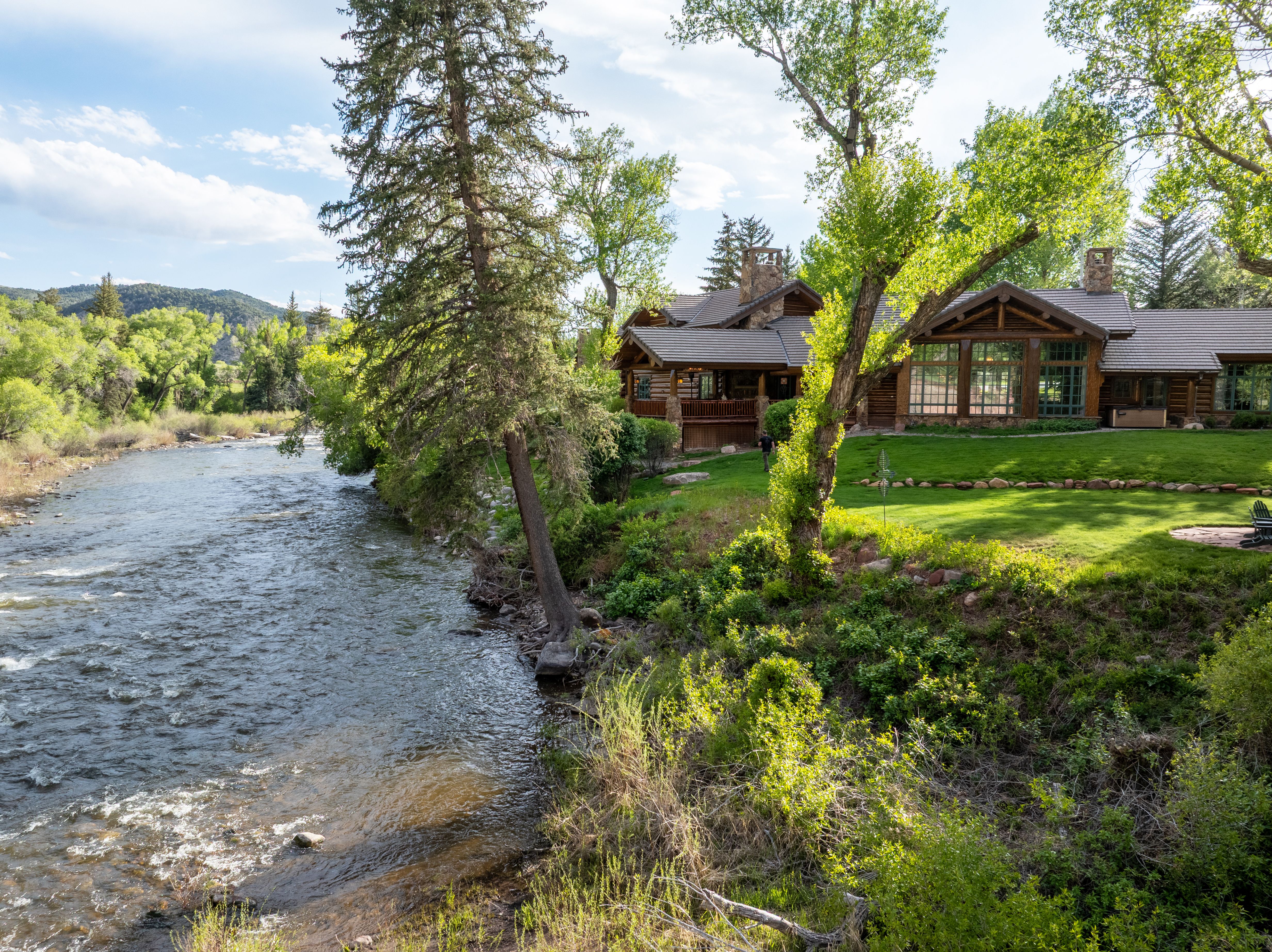 This stunning log cabin estate is nestled along the banks of a rushing river, offering a serene and picturesque setting. The home features classic rustic architecture with large windows that capture the natural light and expansive views of the surrounding lush greenery and mountains. A manicured lawn leads directly to the water's edge, creating a perfect blend of luxury living and natural beauty.