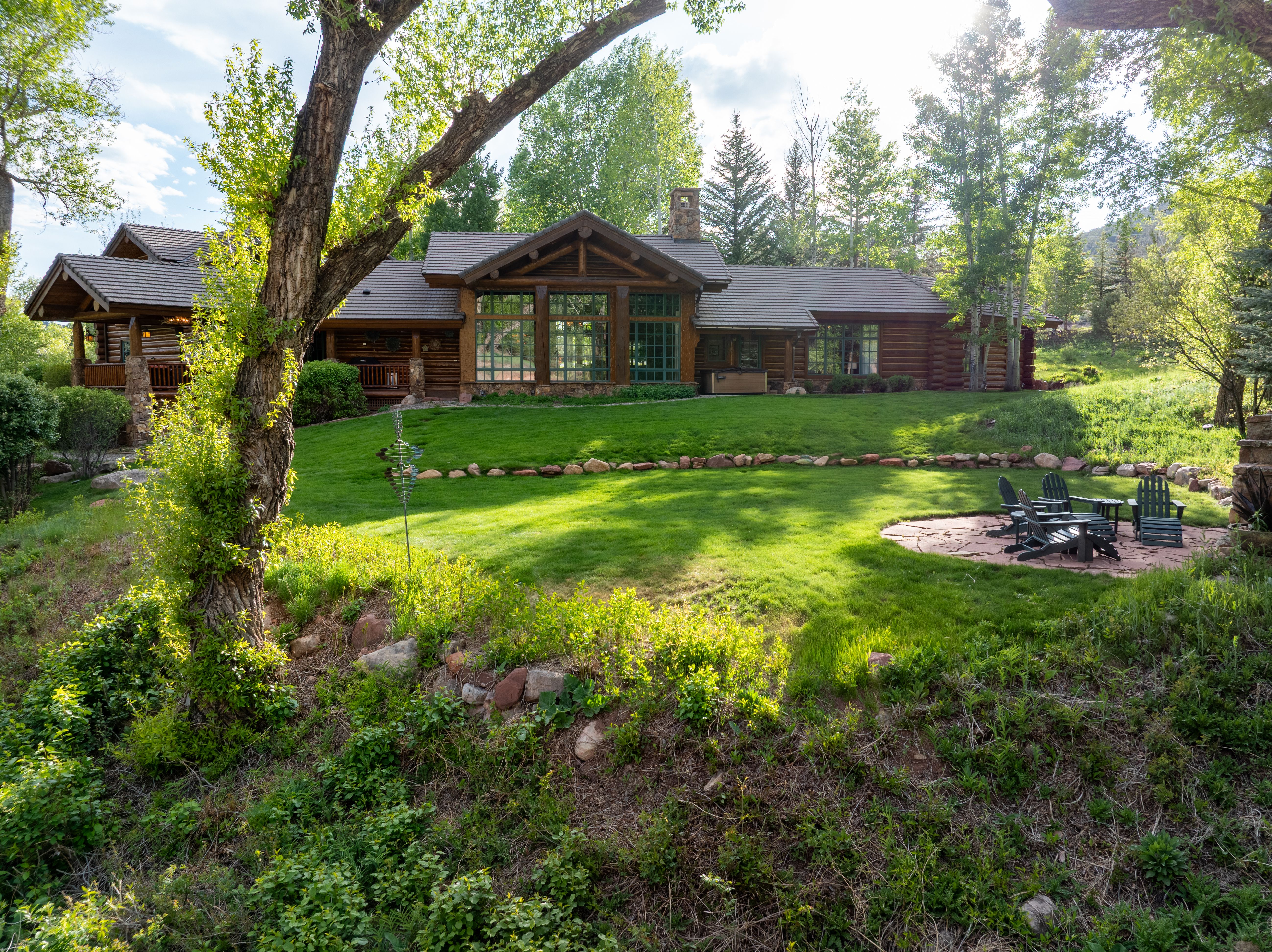 This image showcases the rear exterior of a rustic log cabin-style home, featuring expansive windows and a stone chimney set against a lush, green landscape. A stone patio with Adirondack chairs sits on a manicured lawn, framed by mature trees and natural vegetation. The perspective is a wide, eye-level shot that captures the serene and secluded atmosphere of the property.