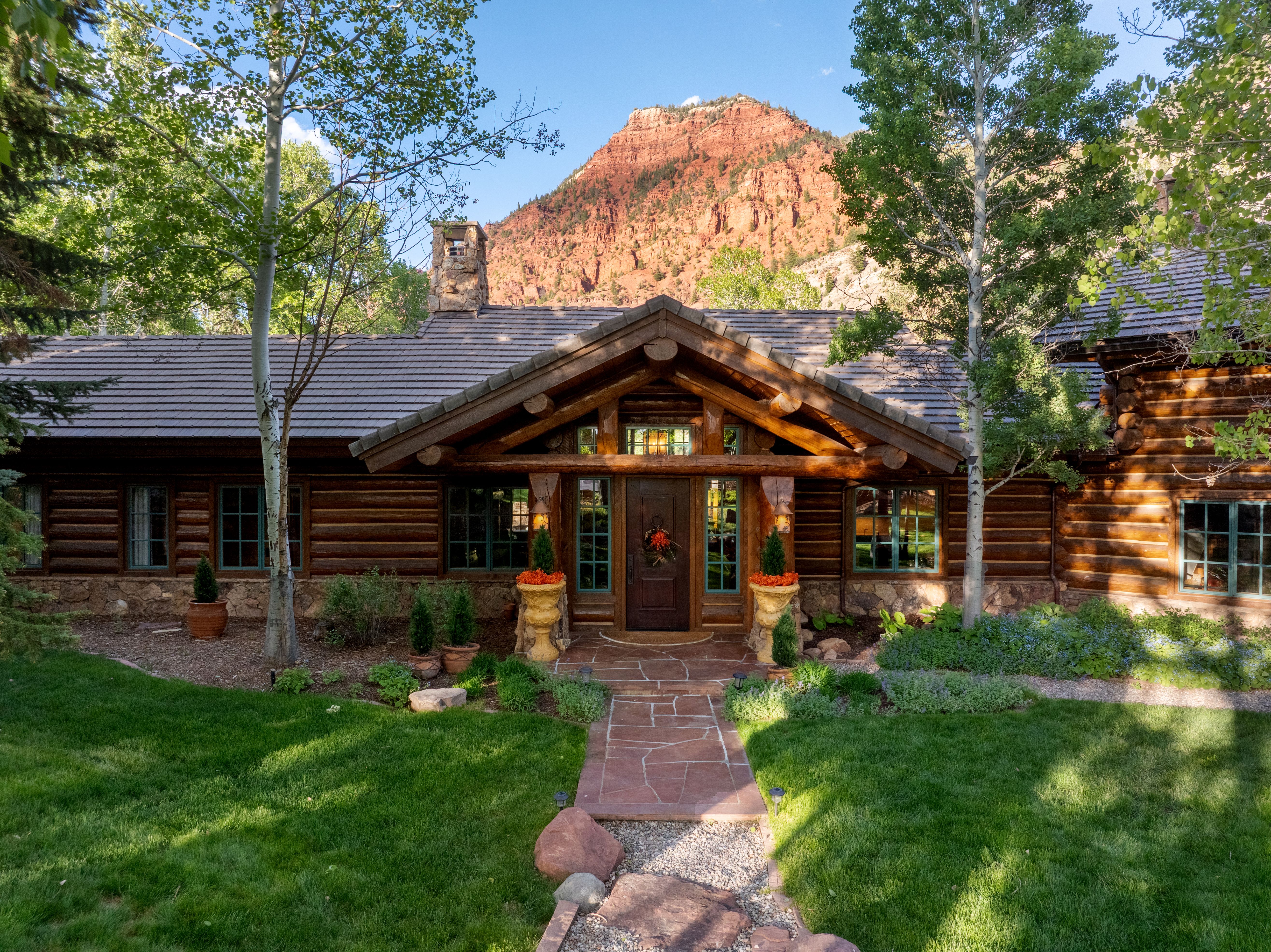 This stunning log cabin home features a rustic, high-end aesthetic with natural stone accents and a prominent gabled entryway. The front facade is framed by lush green lawns and mature aspen trees, with a dramatic red rock mountain serving as a breathtaking backdrop. The stone pathway leads directly to the inviting front door, creating a warm and welcoming atmosphere that highlights the property's serene, mountainous setting.