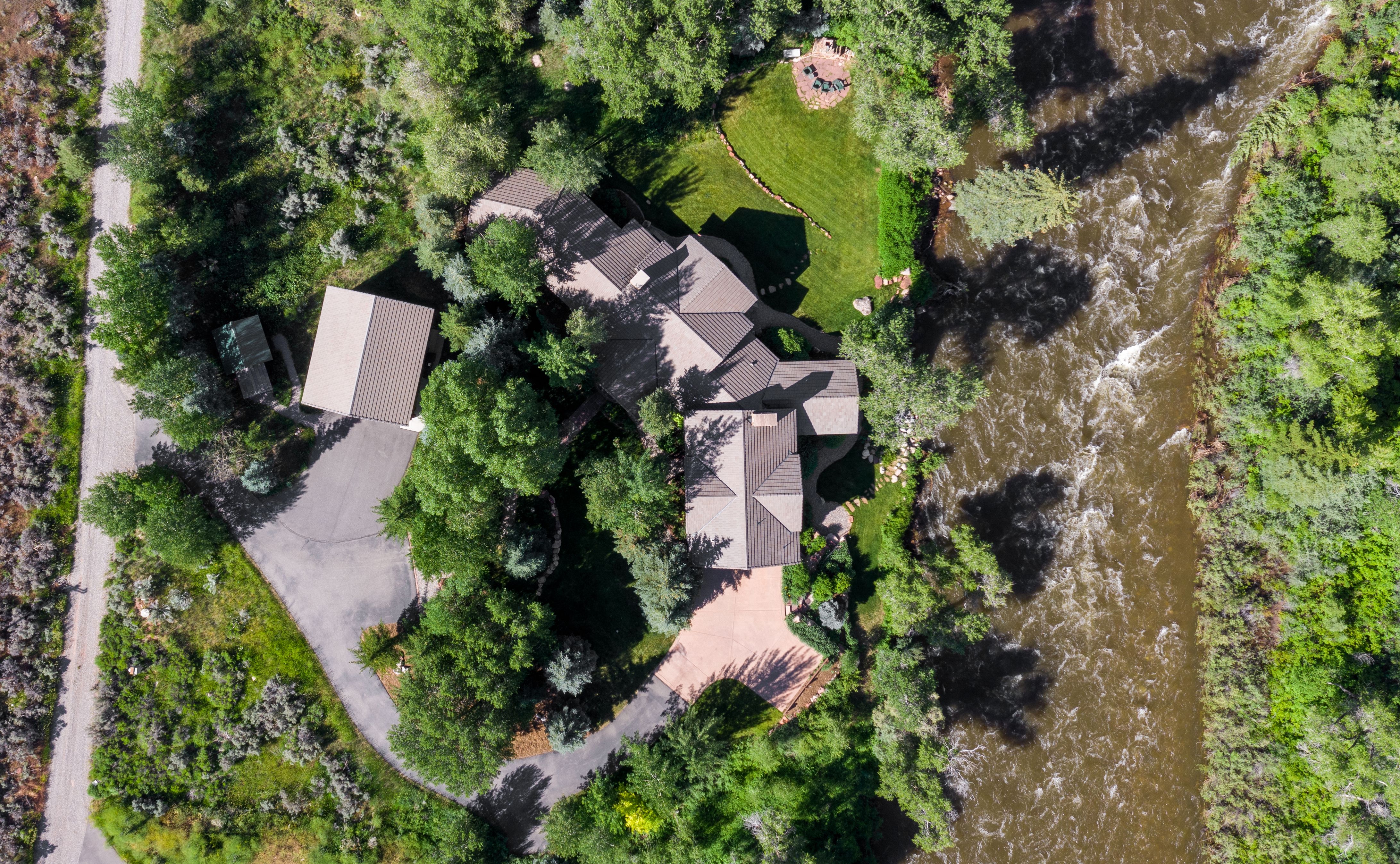 This high-angle aerial perspective captures a secluded residential property nestled along the banks of a rushing river. The home features a multi-gabled roofline, a paved driveway leading to a detached garage, and a lush, tree-lined landscape that provides significant privacy. The setting offers a serene, natural aesthetic, highlighting the seamless integration between the architecture and the surrounding wilderness.
