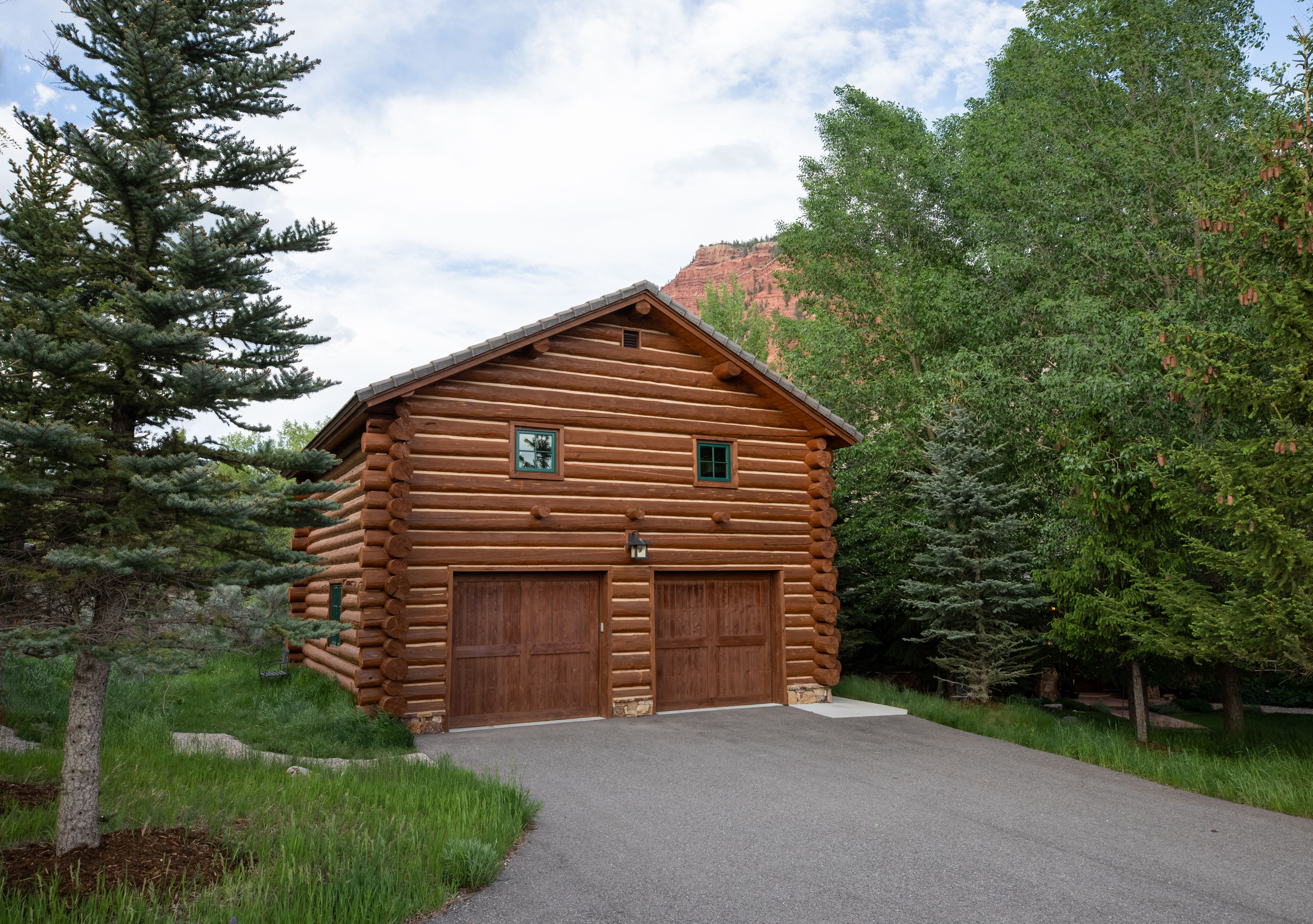 This image showcases a rustic, two-story log cabin-style garage nestled in a lush, wooded landscape. The structure features dark wood logs, two matching garage doors, and two small windows on the upper level, all set against a backdrop of vibrant green trees and a distant red rock mountain. The perspective is a ground-level shot from the paved driveway, emphasizing the home's integration with its natural, serene environment.