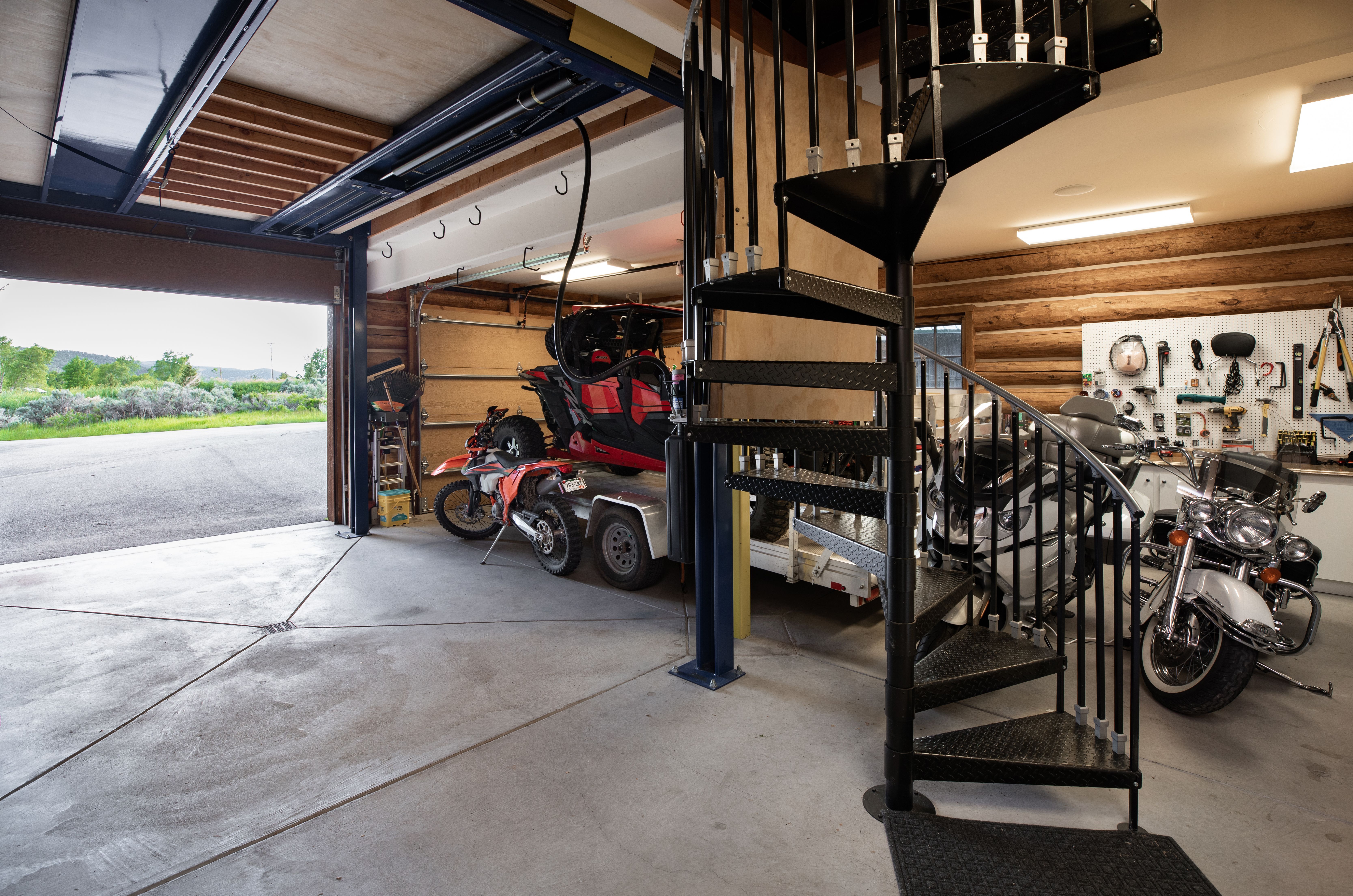 This spacious, rustic-style garage features log walls and a concrete floor, housing a variety of recreational vehicles including a UTV, a dirt bike, and two motorcycles. A prominent black metal spiral staircase serves as a central architectural feature, connecting the ground level to an upper floor. The open garage door reveals a scenic view of the surrounding landscape, emphasizing the property's outdoor-oriented lifestyle.