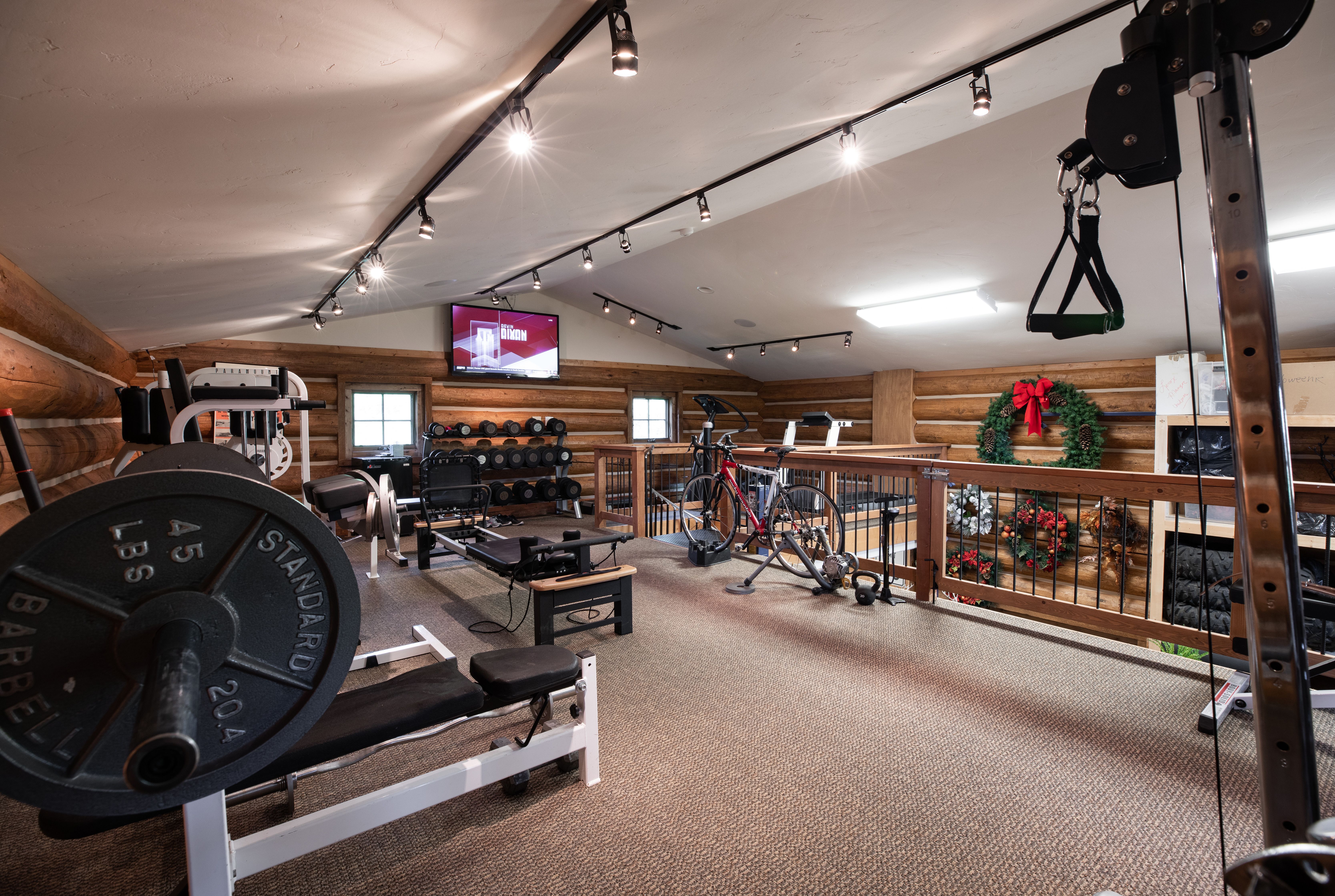 This spacious home gym is situated in a rustic loft area with exposed log walls and a vaulted ceiling. The room is well-equipped with a variety of fitness gear, including a weight bench, a rowing machine, a stationary bike, and a rack of dumbbells. A television is mounted on the wall, and the space overlooks a lower level, creating an open and functional workout environment.