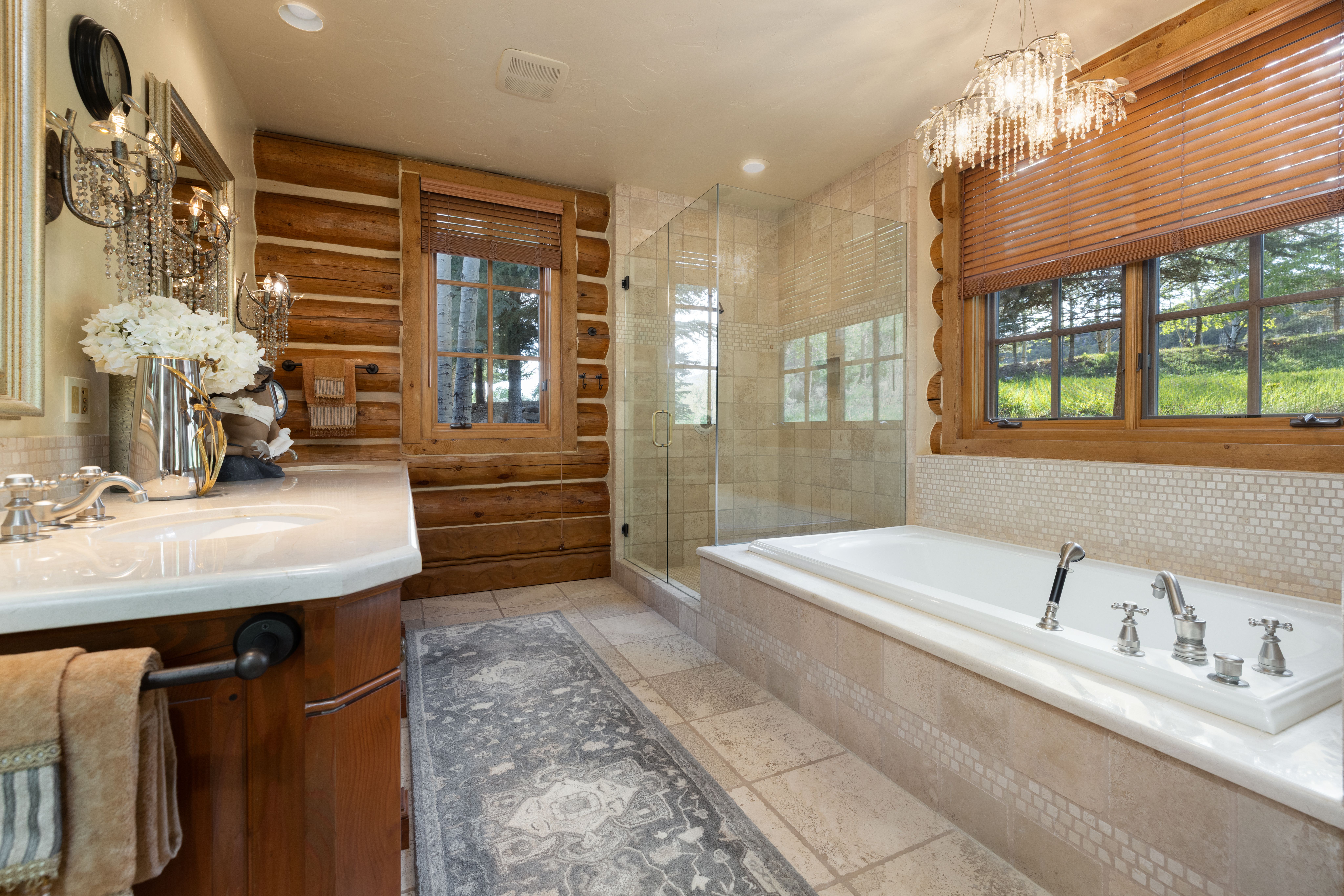 This luxurious primary bathroom features a rustic log cabin aesthetic combined with elegant, high-end finishes. The space includes a deep soaking tub set against a tiled surround, a glass-enclosed walk-in shower, and a vanity with a stone countertop, all complemented by a crystal chandelier and warm wood walls. The perspective captures the entire room, highlighting the harmonious blend of natural textures and sophisticated lighting.