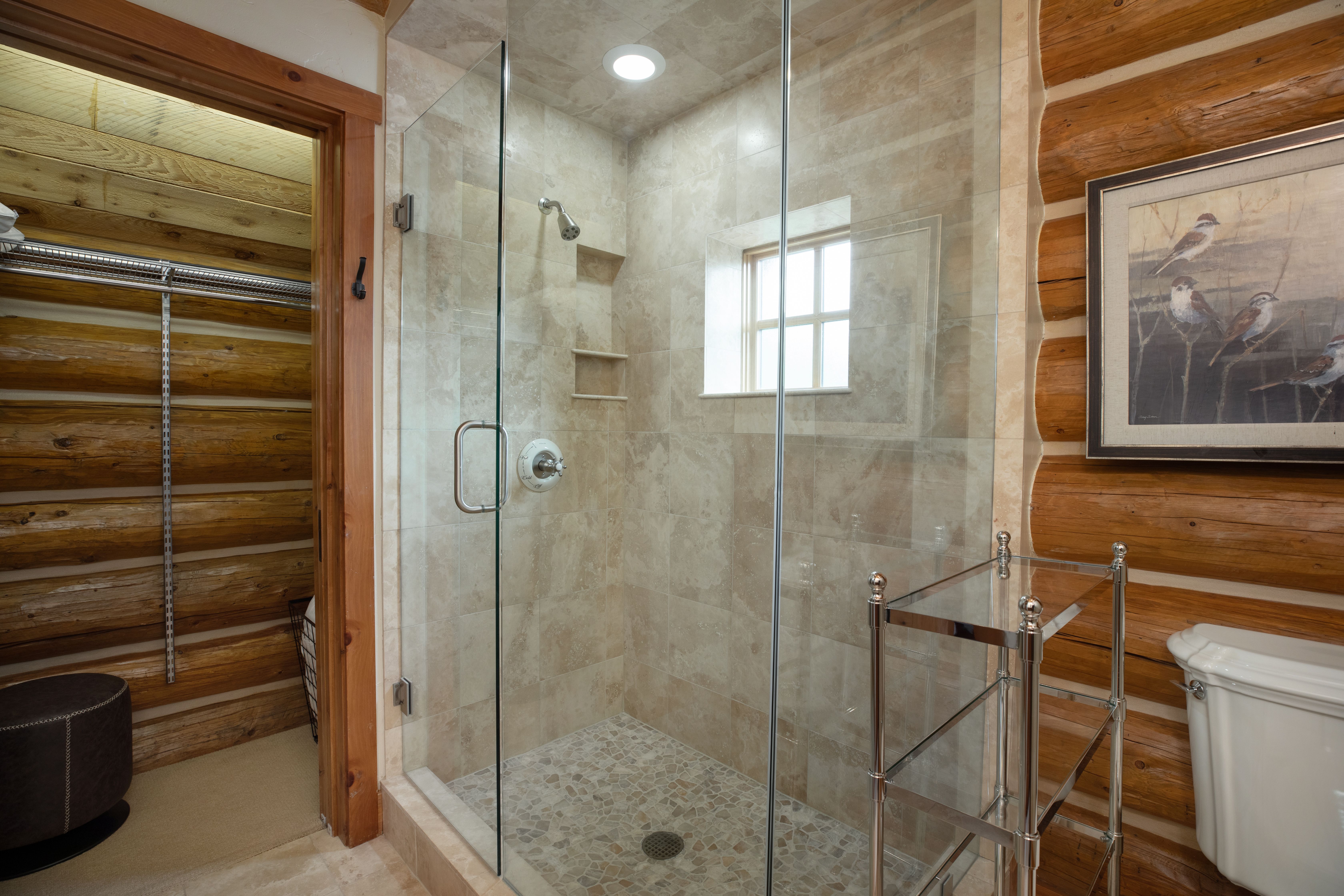 This bathroom features a rustic log cabin aesthetic, highlighted by exposed timber walls and a walk-in shower with stone tile flooring and walls. A glass-enclosed shower stall sits adjacent to a chrome shelving unit and a white toilet, while a doorway leads into a walk-in closet with matching log walls. The space combines natural, rugged textures with modern bathroom fixtures for a cozy, high-end mountain retreat feel.