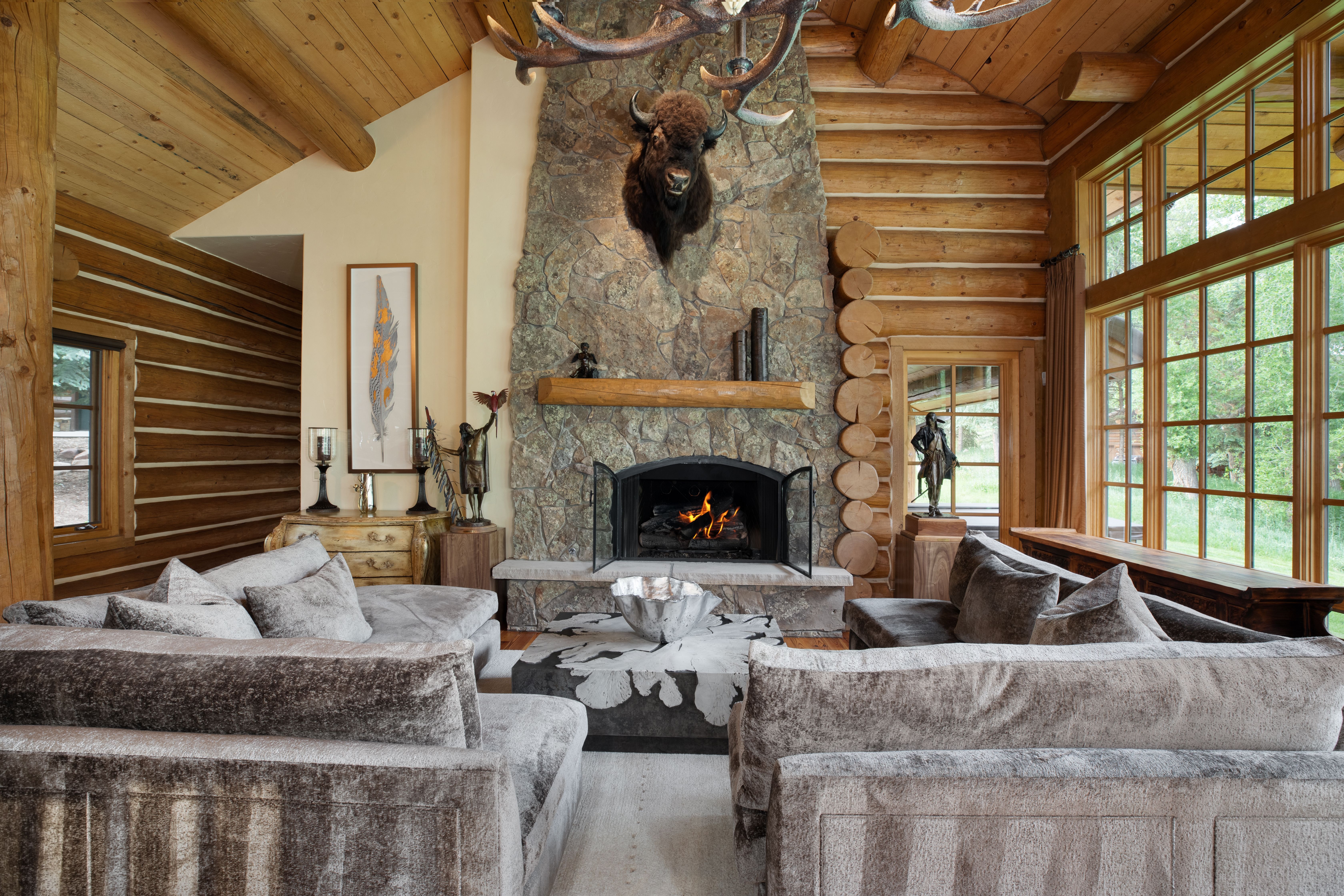 This rustic living room features a grand stone fireplace as its focal point, adorned with a mounted bison head and antler chandelier. The space is framed by warm log walls and expansive windows that invite natural light and views of the outdoors. Plush, textured grey sofas are arranged around a unique, modern coffee table, creating a cozy and inviting atmosphere perfect for a mountain retreat.