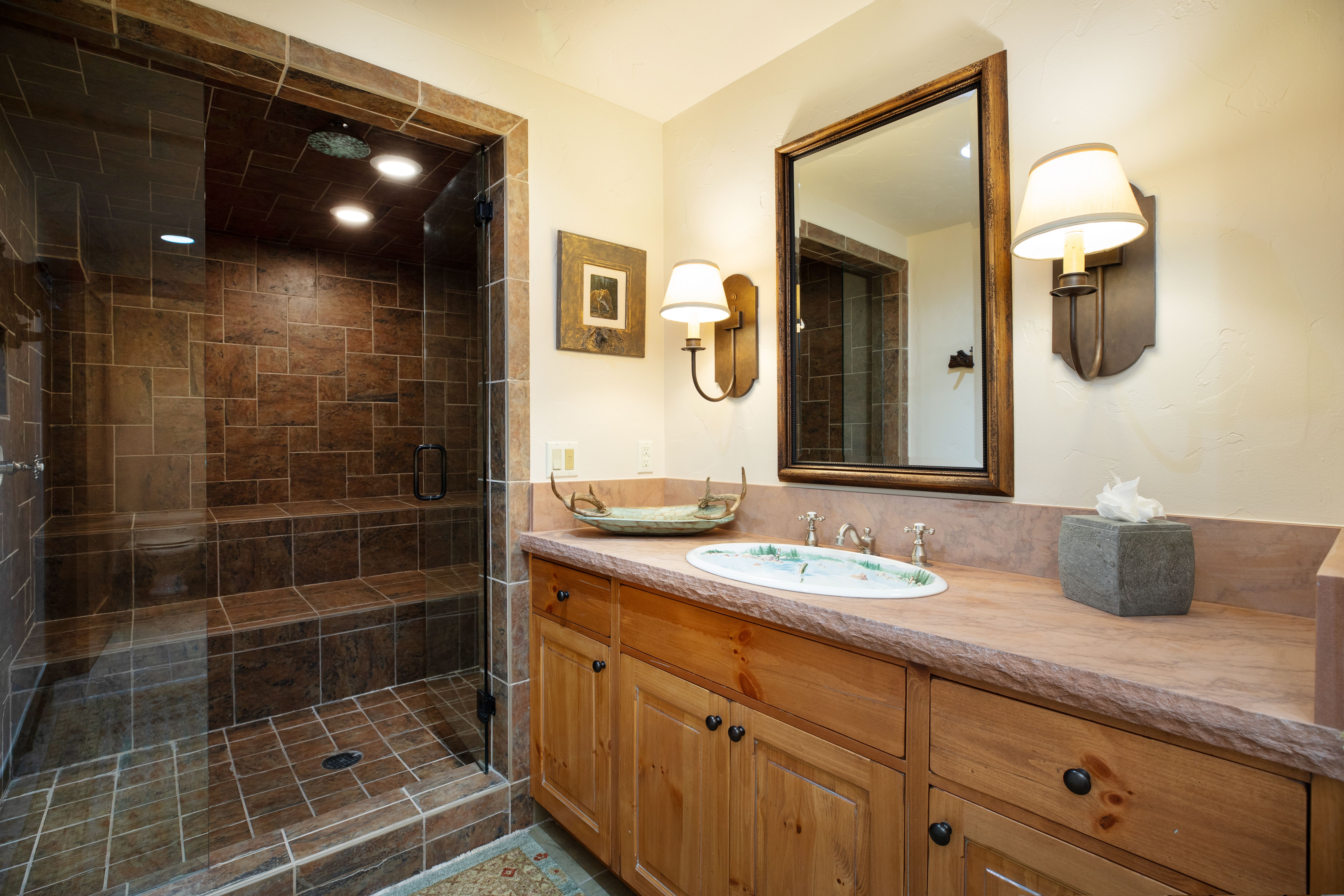 This rustic-style bathroom features a custom stone-topped vanity with a unique painted sink and a spacious walk-in steam shower enclosed by glass. The warm wood cabinetry, antler-themed decor, and rich earth-toned tile work create a cozy, lodge-inspired atmosphere. The perspective is a straight-on shot of the vanity and shower area, highlighting the high-end finishes and thoughtful design details.