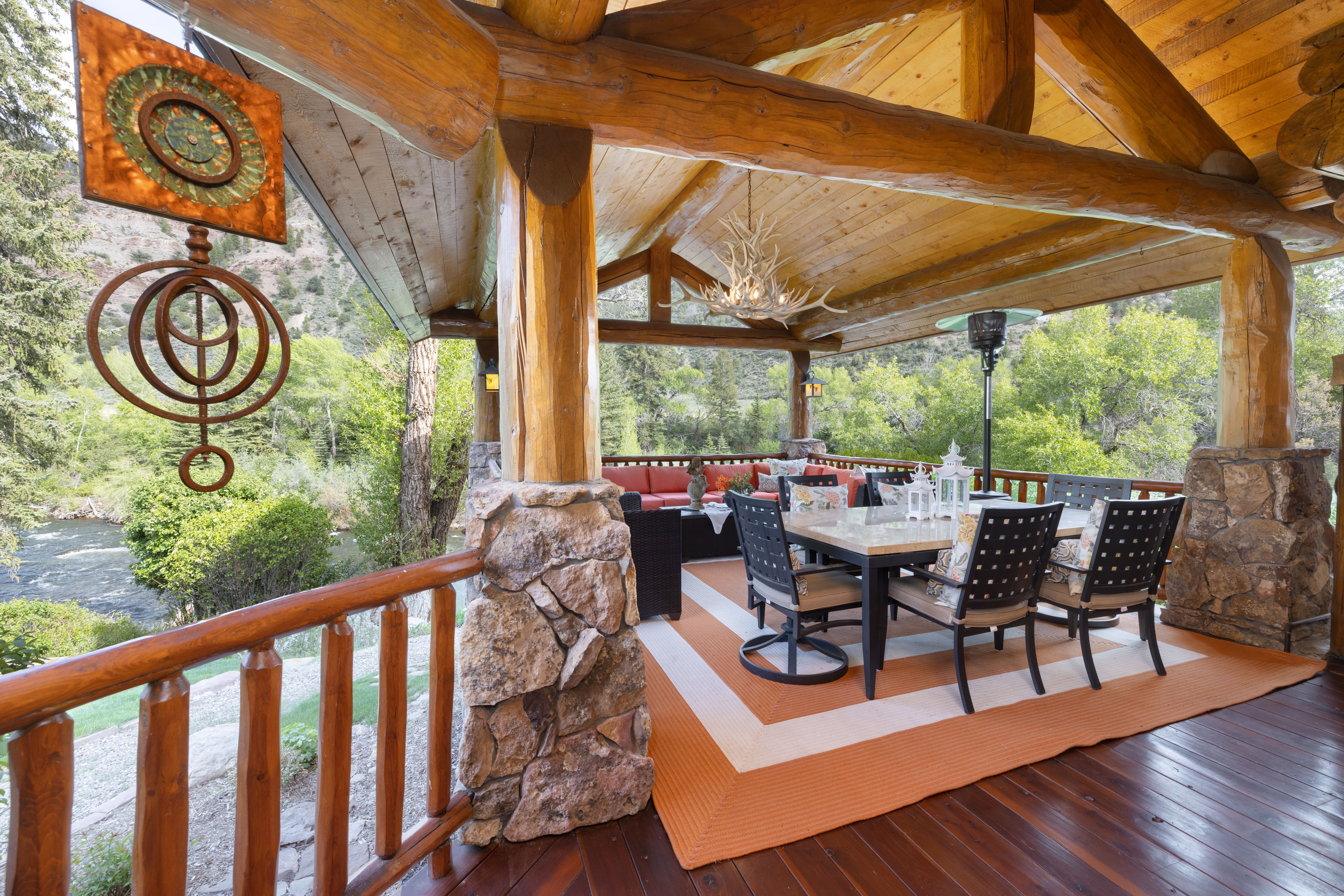 This expansive, rustic covered deck features heavy timber framing, stone-based columns, and a stunning view of a rushing river and lush forest. The space is furnished with a dining set and a comfortable outdoor lounge area, creating an inviting atmosphere for al fresco dining or relaxation. The warm wood tones and natural materials harmonize perfectly with the scenic mountain landscape.
