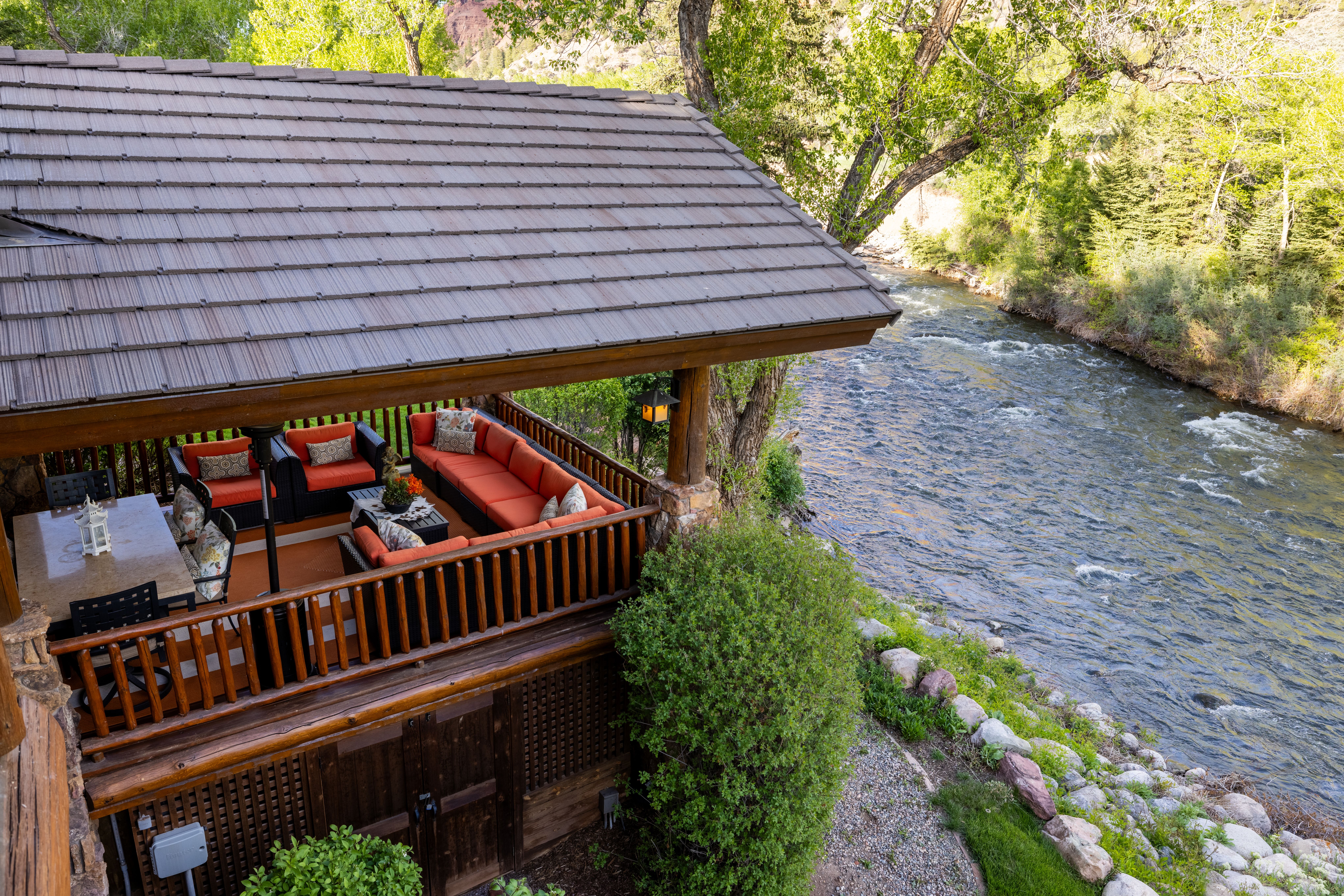 This high-angle shot captures a spacious, covered wooden deck overlooking a rushing river, creating a serene and picturesque outdoor living space. The deck is furnished with comfortable, vibrant orange-cushioned seating and a dining area, all sheltered by a sturdy, tiled roof structure. The surrounding lush greenery and the proximity to the flowing water enhance the tranquil, nature-focused appeal of this property.