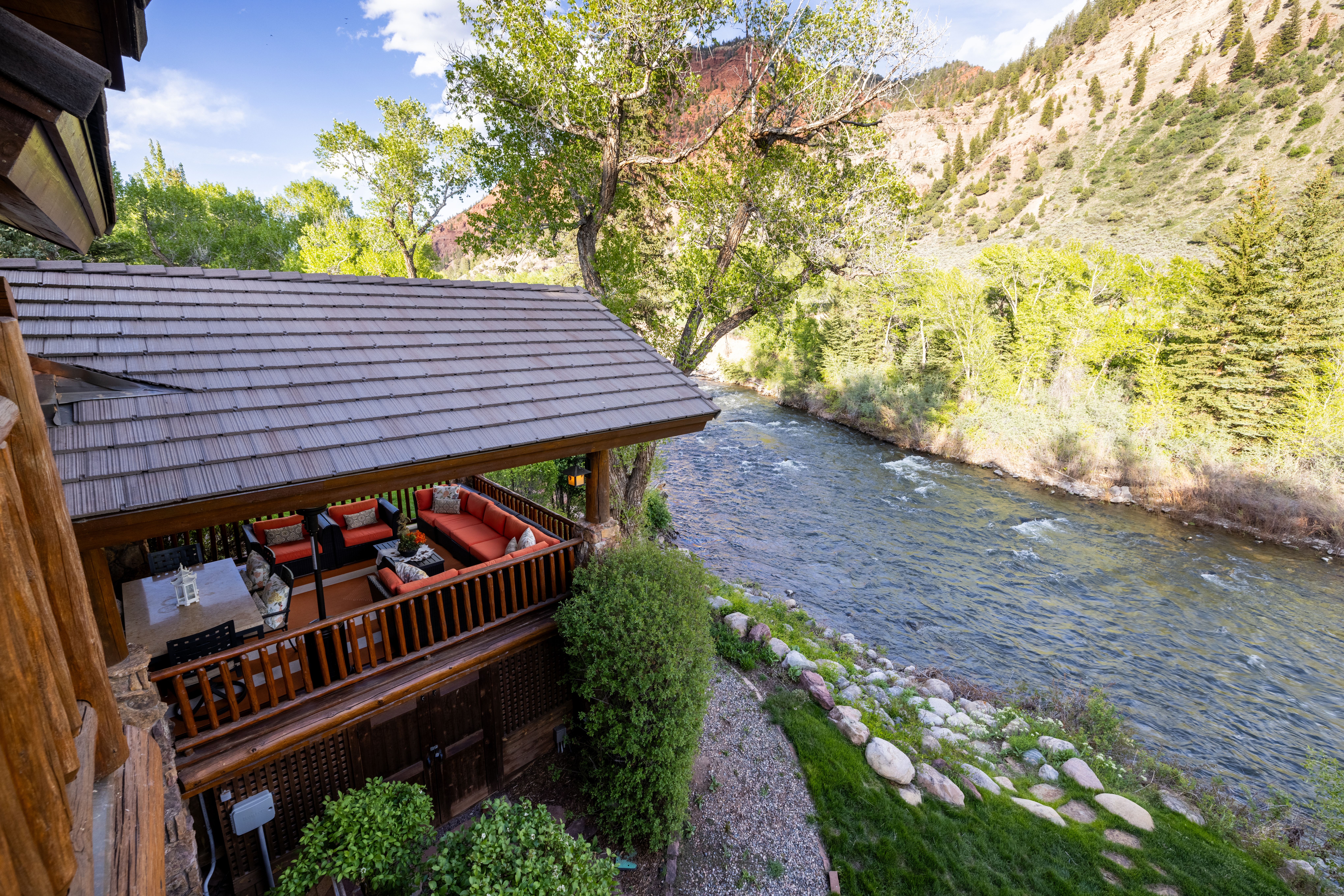 This elevated perspective captures a rustic, covered wooden deck overlooking a scenic, rushing river set against a backdrop of rugged, tree-covered mountains. The deck is furnished with comfortable outdoor seating, including orange-cushioned sofas and a dining table, creating an inviting space for relaxation or entertaining. The natural setting, combined with the warm wood tones of the structure, evokes a peaceful and luxurious mountain retreat atmosphere.