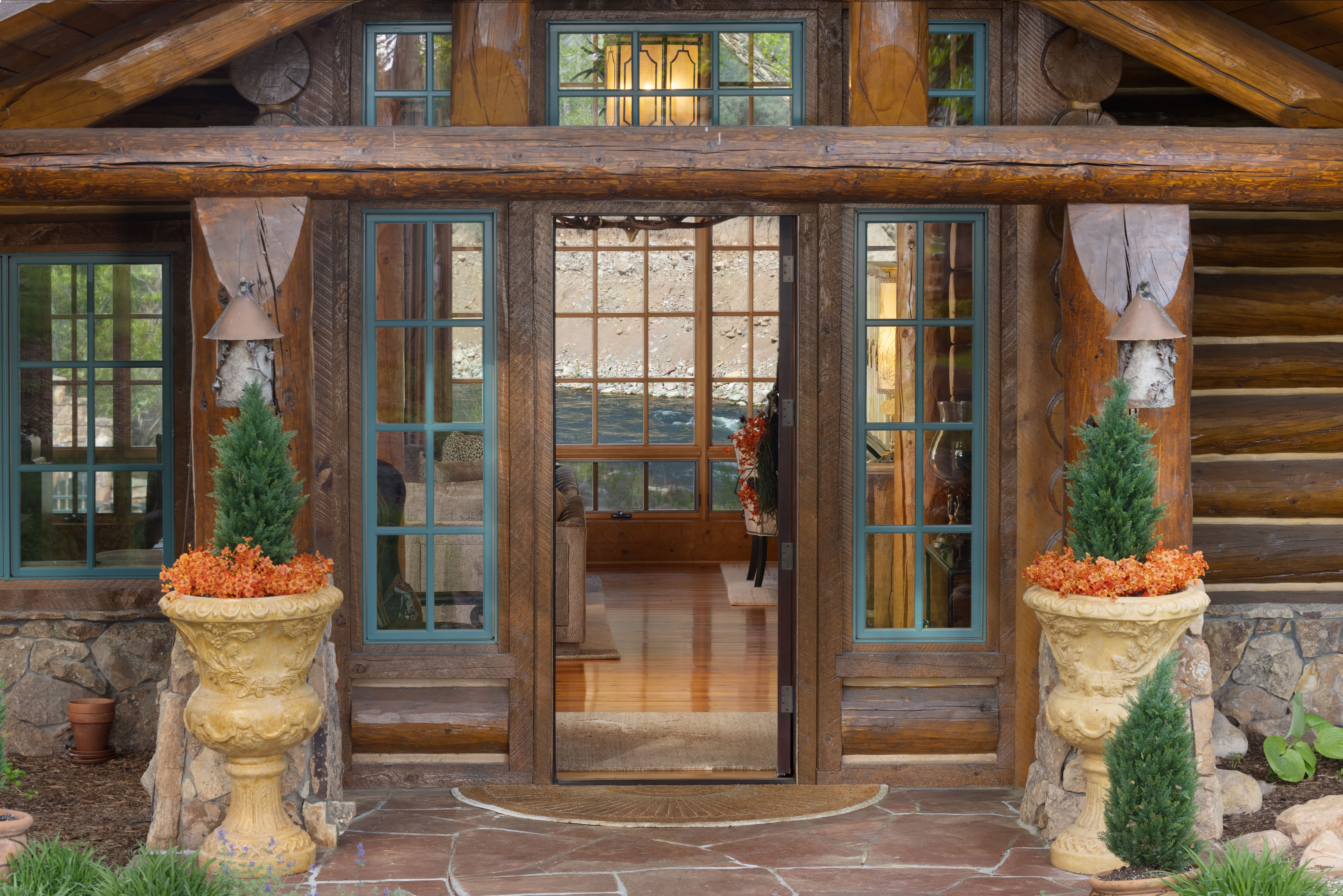 This inviting entryway features a rustic log cabin aesthetic with a prominent timber-framed porch and stone foundation. Symmetrical planters with evergreen shrubs and vibrant orange flowers flank the open doorway, which reveals a warm, polished wood interior and a scenic view of a river beyond. The combination of natural materials and the open-door perspective creates a welcoming and serene atmosphere.