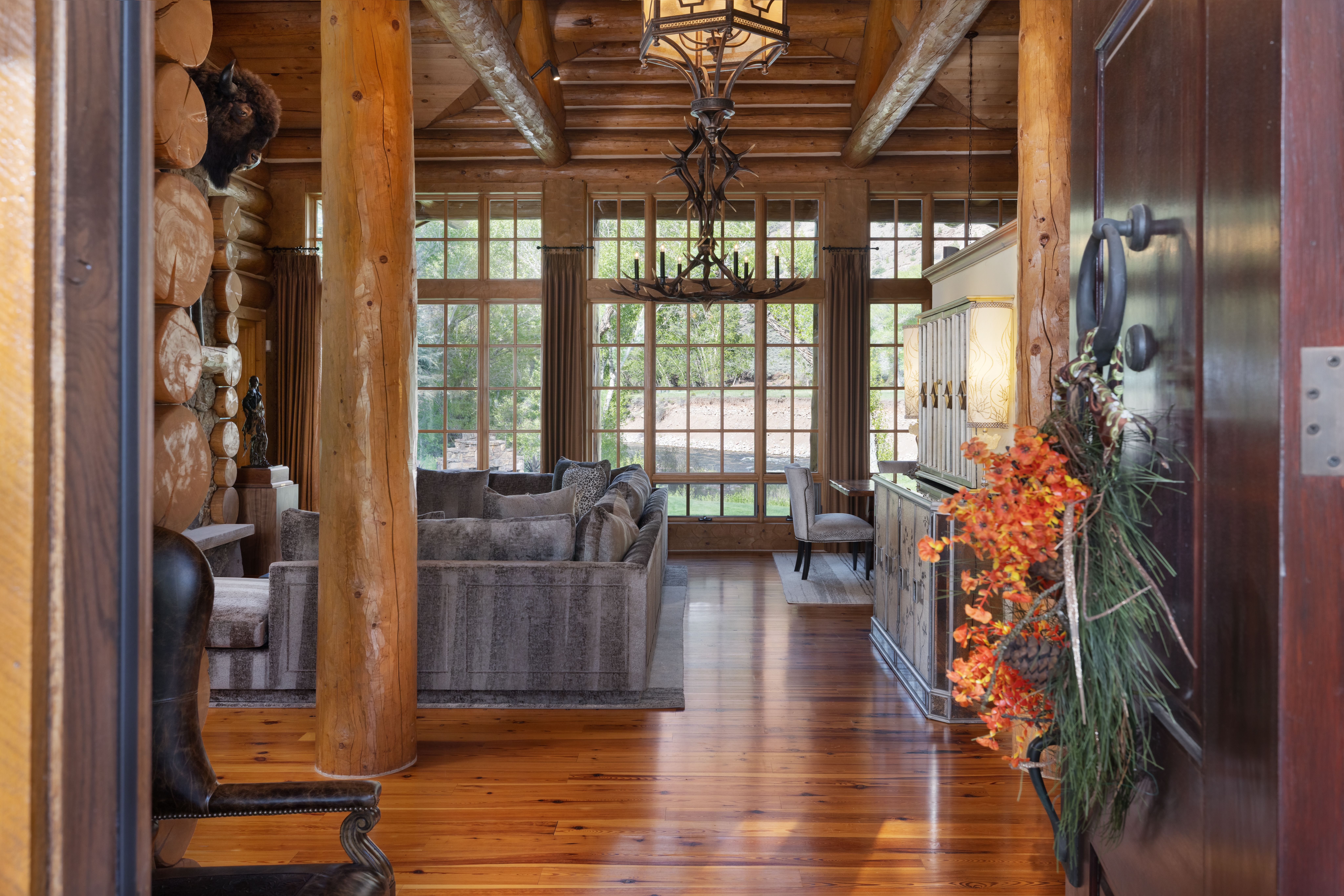 This rustic, high-end living room features massive log walls, exposed timber ceiling beams, and polished hardwood floors, creating a warm and inviting mountain lodge aesthetic. A large, plush grey sectional sofa anchors the space, while a striking antler chandelier hangs above, drawing the eye toward the expansive floor-to-ceiling windows that frame a serene outdoor view. The perspective is captured from an entryway, offering a cinematic transition into the open-concept living area.