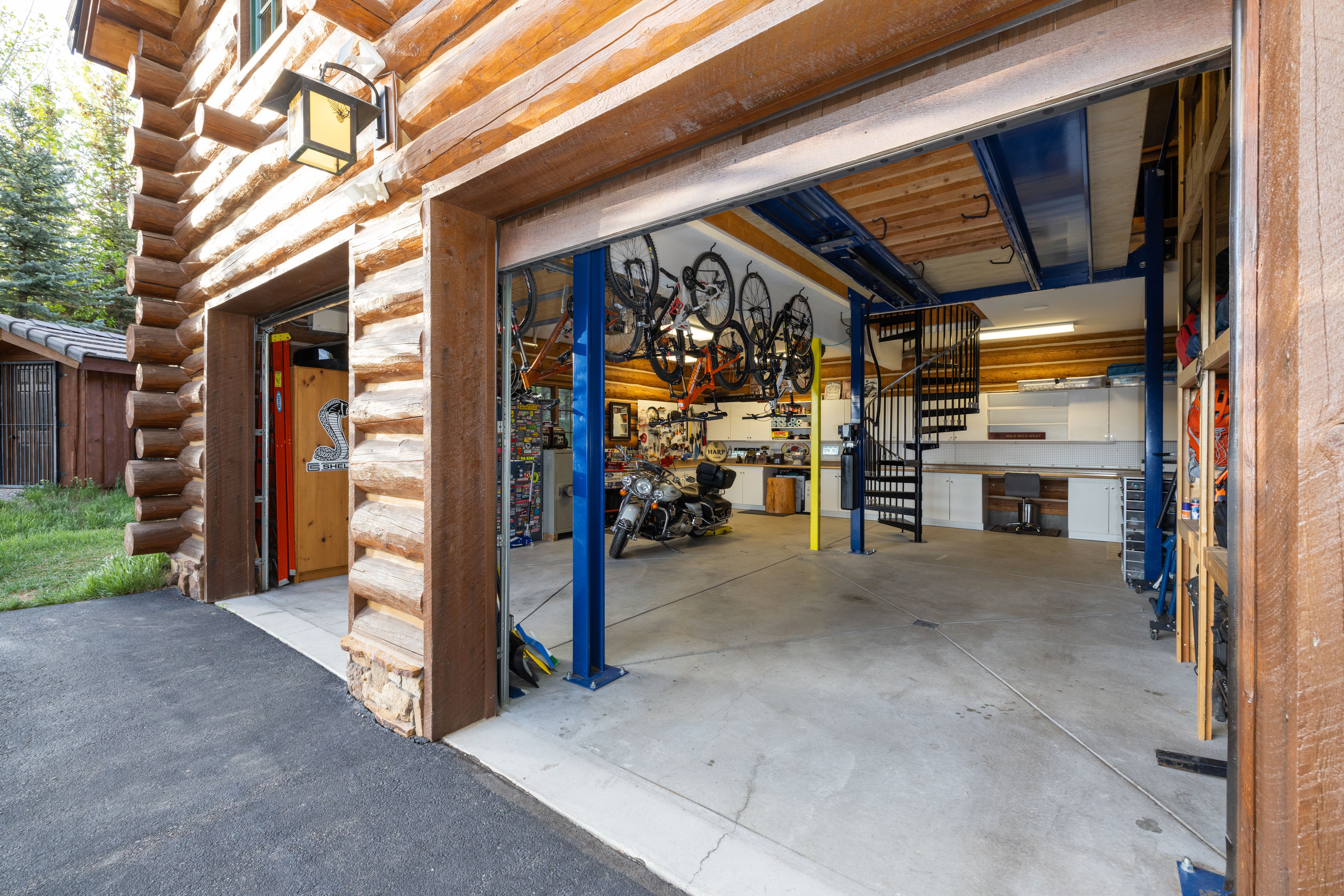 This image showcases a spacious, rustic-style garage built with log cabin architecture, featuring an open bay that reveals a well-organized workshop area. The interior includes a motorcycle, multiple bicycles suspended from the ceiling, a spiral staircase, and a dedicated workspace with cabinetry. The perspective is from the driveway looking into the garage, highlighting the blend of rugged exterior aesthetics and a highly functional, modern interior space.
