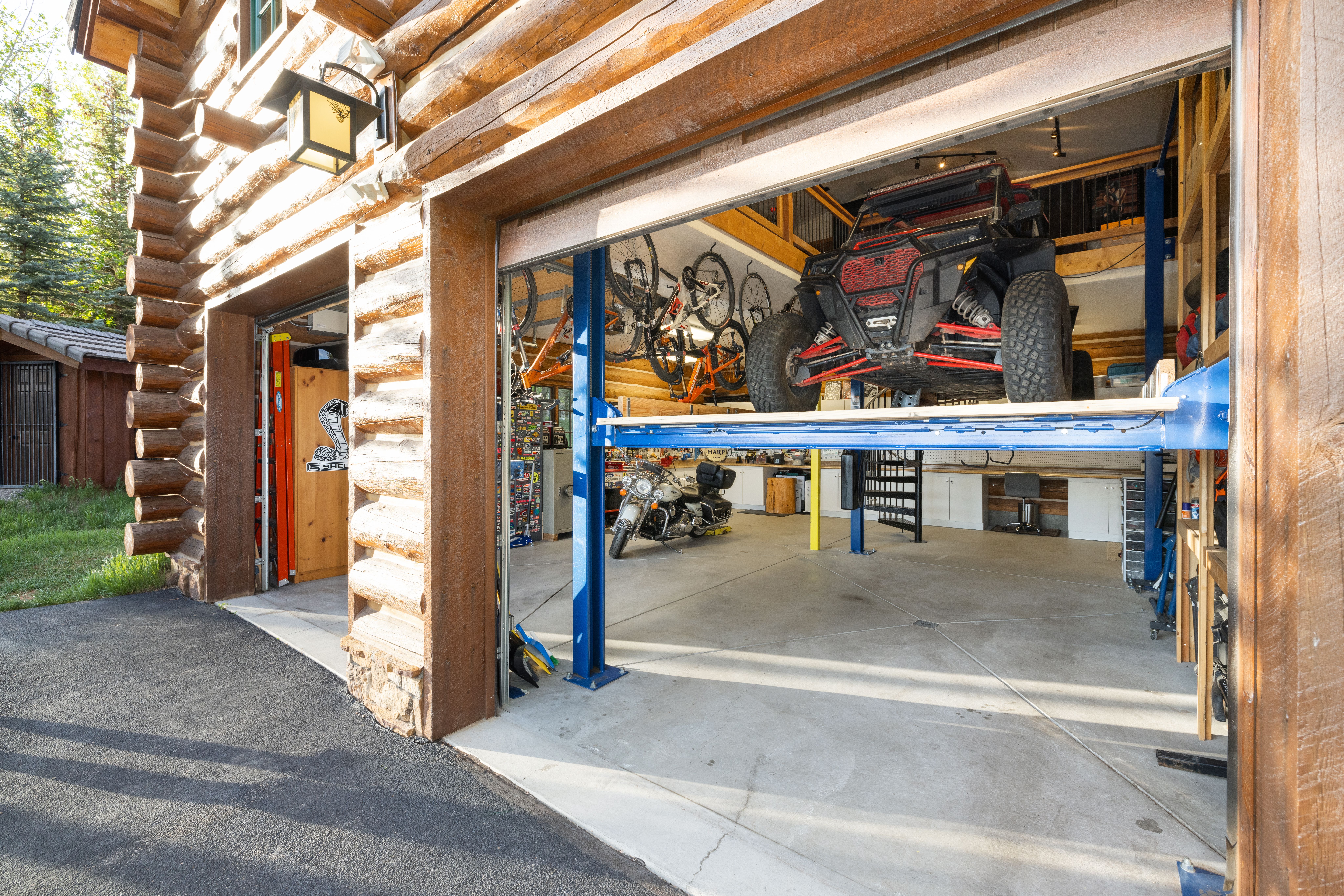 This image captures a spacious, rustic log-cabin style garage with an open bay revealing a professional-grade vehicle lift holding an off-road utility vehicle. The interior is well-organized, featuring a motorcycle, wall-mounted bicycles, and a clean concrete floor, suggesting a high-end workshop or hobby space. The perspective from the driveway highlights the unique architectural character of the log construction and the functional, multi-level storage capabilities of the garage.