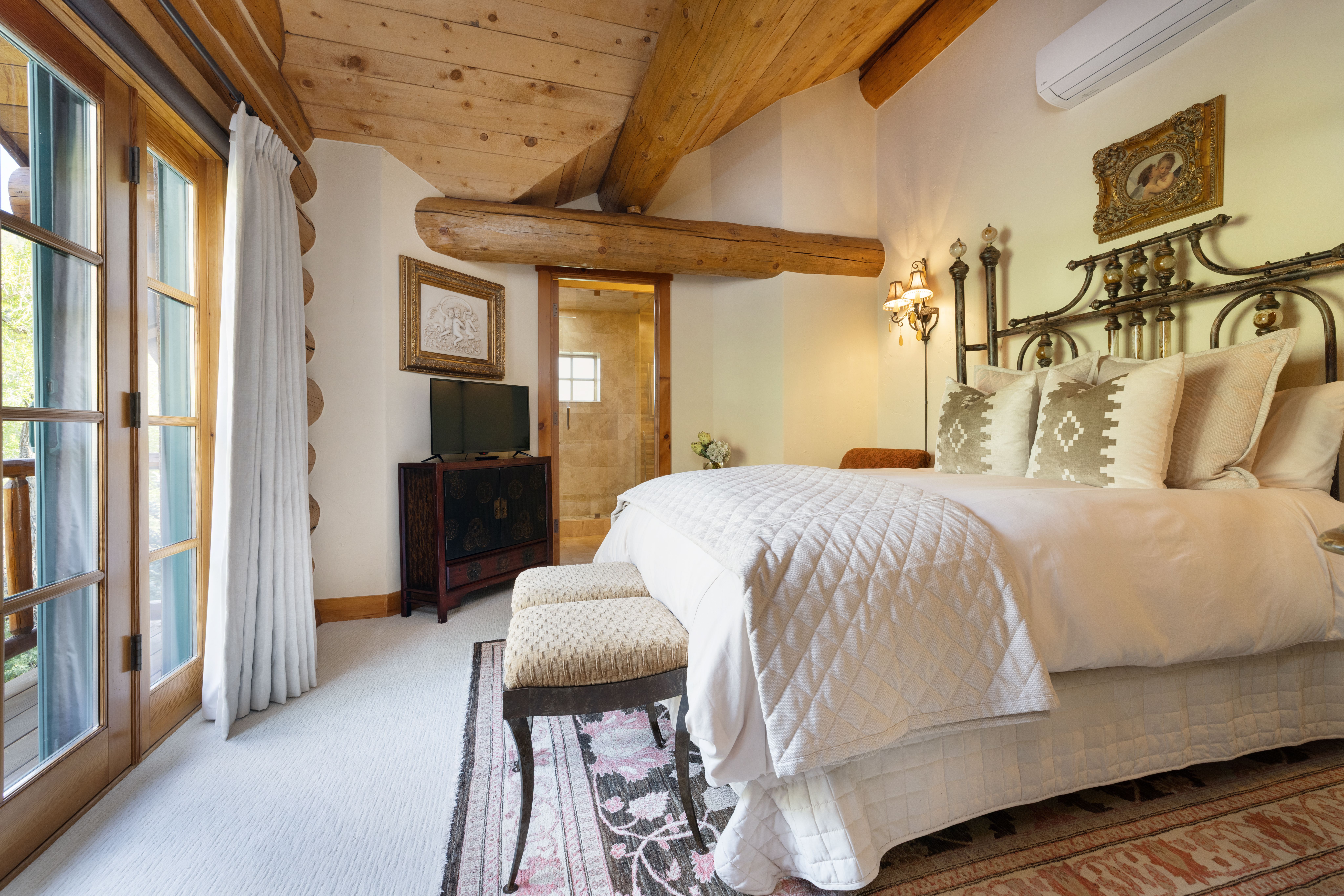 This cozy primary bedroom features a rustic log-cabin aesthetic with exposed timber ceiling beams and warm, neutral tones. The room is anchored by a large bed with a decorative metal headboard, flanked by elegant wall sconces, and includes a private entrance to a balcony through glass doors. A small media console and a doorway leading to an en-suite bathroom complete this inviting and well-appointed space.