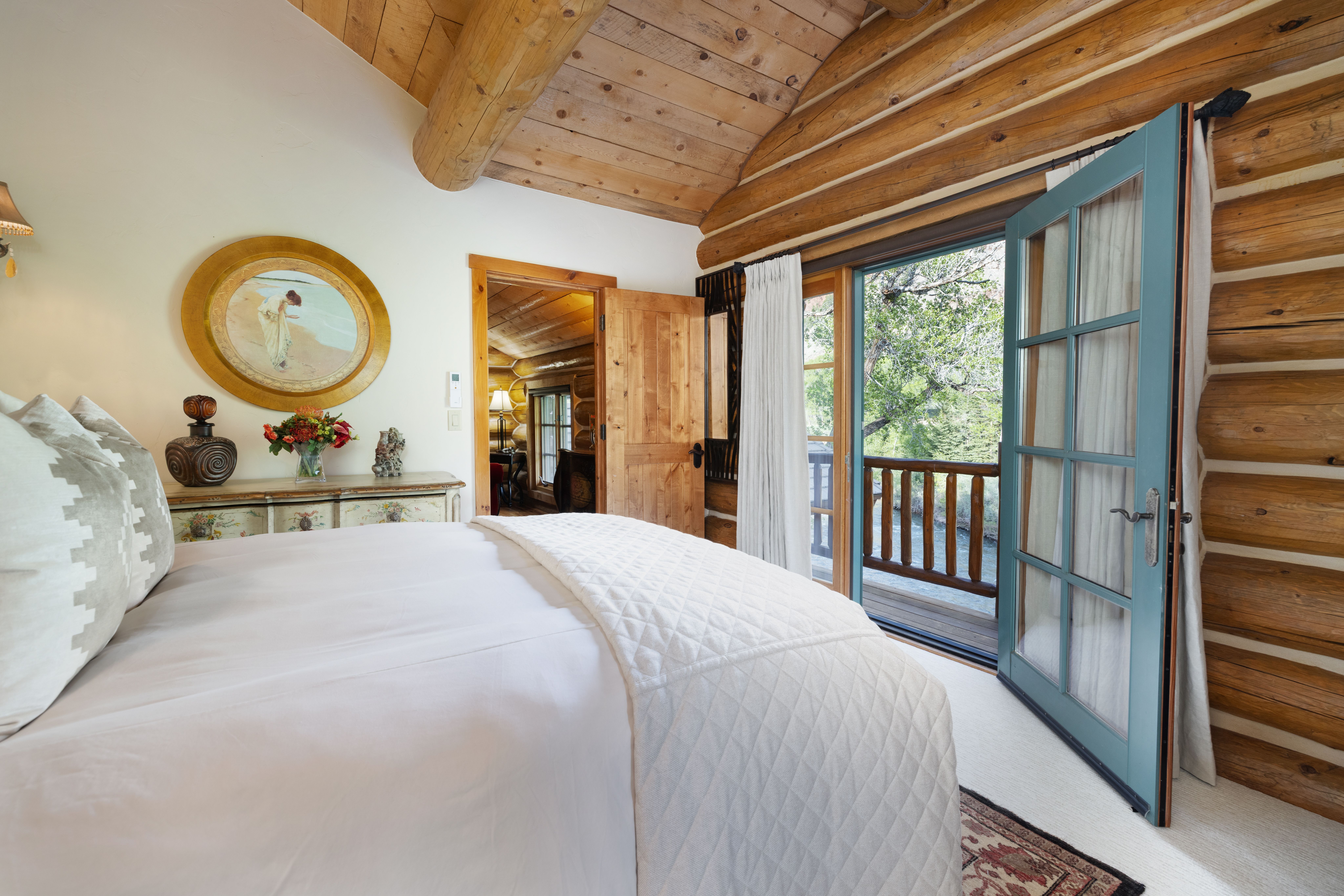 This cozy primary bedroom features rustic log walls and a vaulted wood-paneled ceiling, creating a warm, cabin-like atmosphere. A plush bed with white bedding sits prominently in the foreground, while a blue-framed glass door opens onto a private balcony with lush forest views. The room is accented by a circular framed painting and a decorative dresser, blending traditional mountain charm with comfortable living.