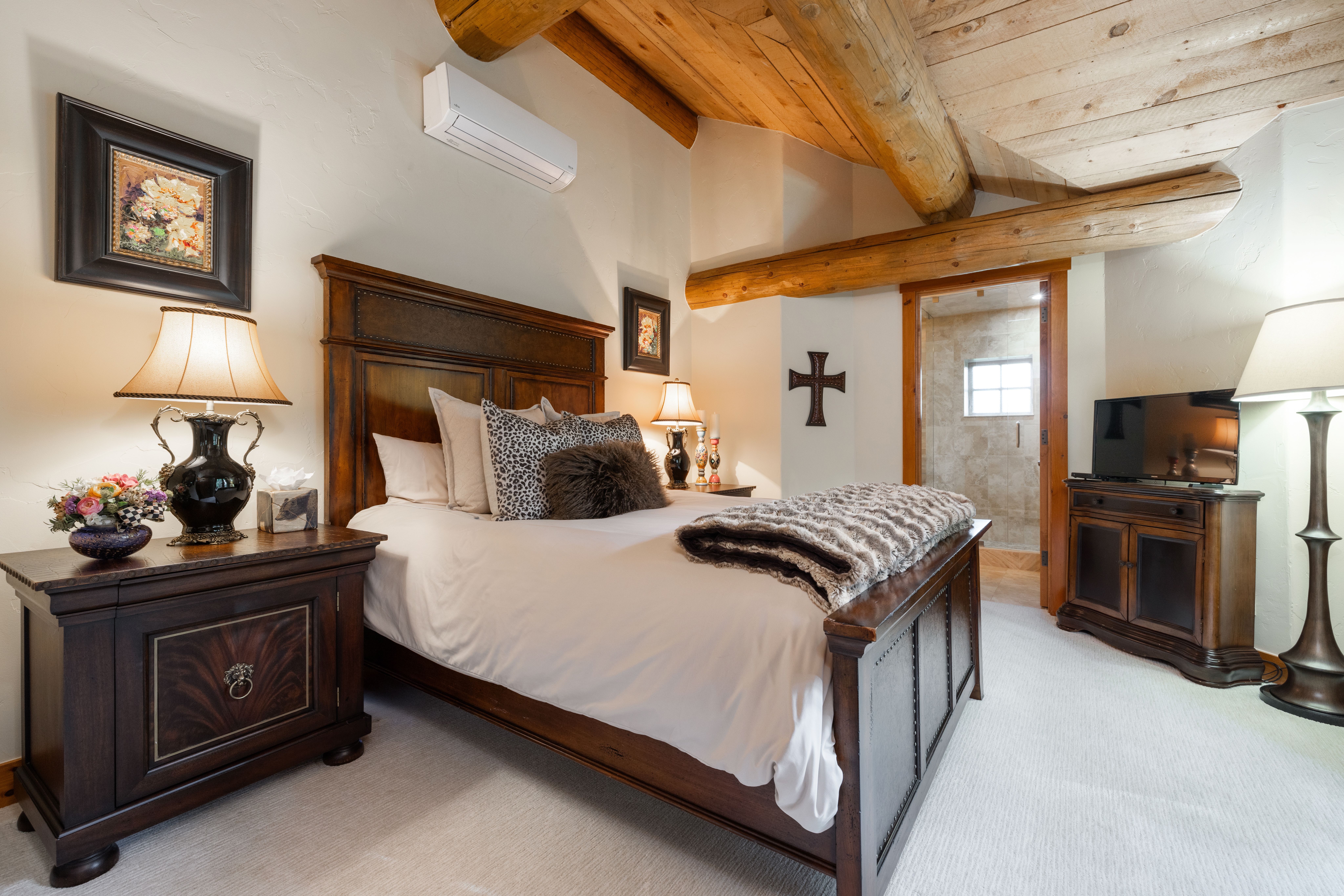 This cozy primary bedroom features a rustic aesthetic with exposed wooden ceiling beams and a warm, inviting atmosphere. The room is anchored by a substantial dark wood bed frame, complemented by matching nightstands, a television console, and a plush, neutral-toned carpet. A doorway leads into an adjacent tiled bathroom, while a wall-mounted air conditioning unit and a decorative cross add functional and stylistic touches to the space.