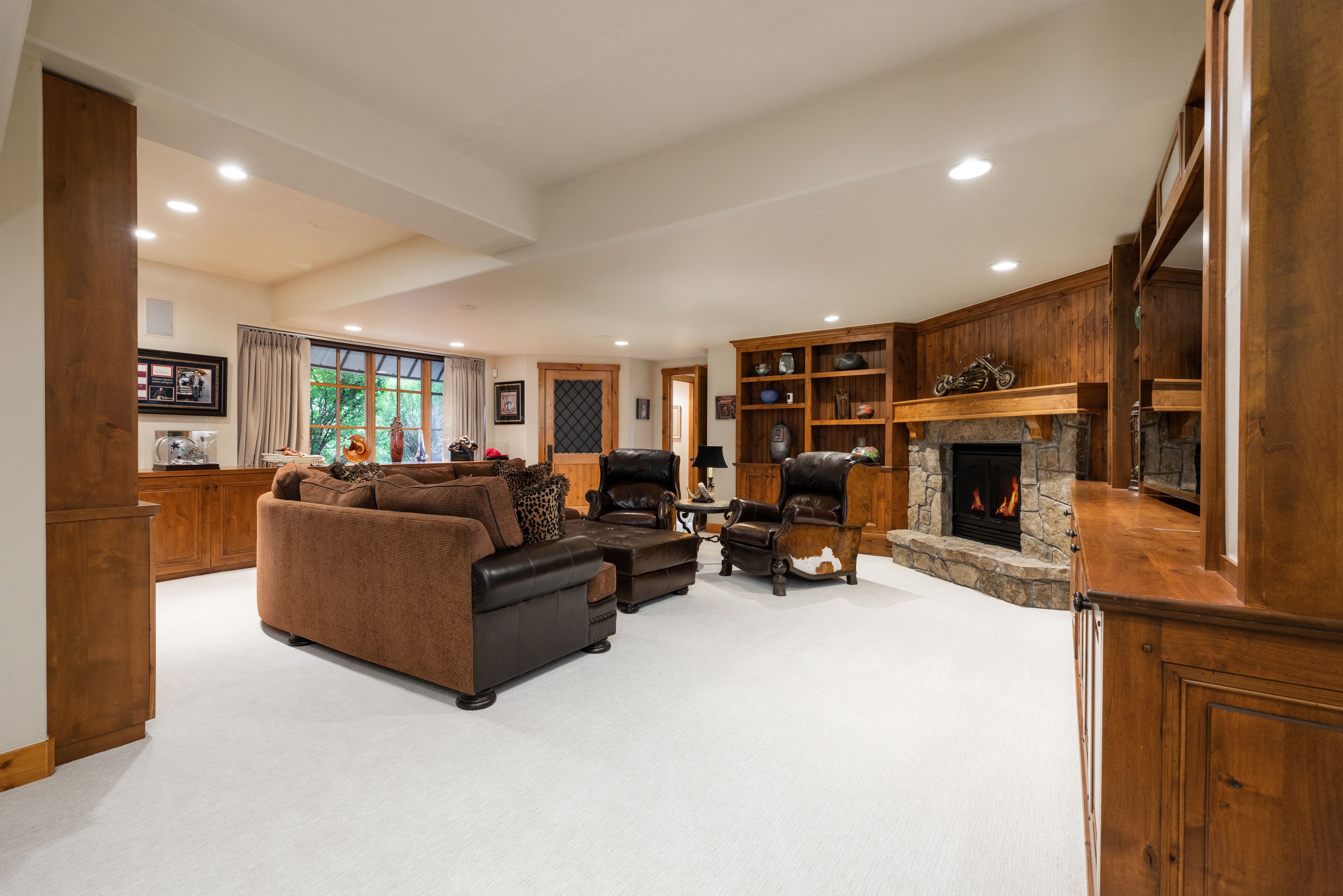 This cozy, rustic-style basement living area features a warm color palette with rich wood paneling, built-in shelving, and a stone fireplace. The space is furnished with a comfortable brown sectional sofa, leather armchairs, and a matching ottoman, all set against a clean, light-colored carpet. The perspective is a wide-angle shot from the corner of the room, capturing the inviting atmosphere and the seamless integration of the fireplace and entertainment area.