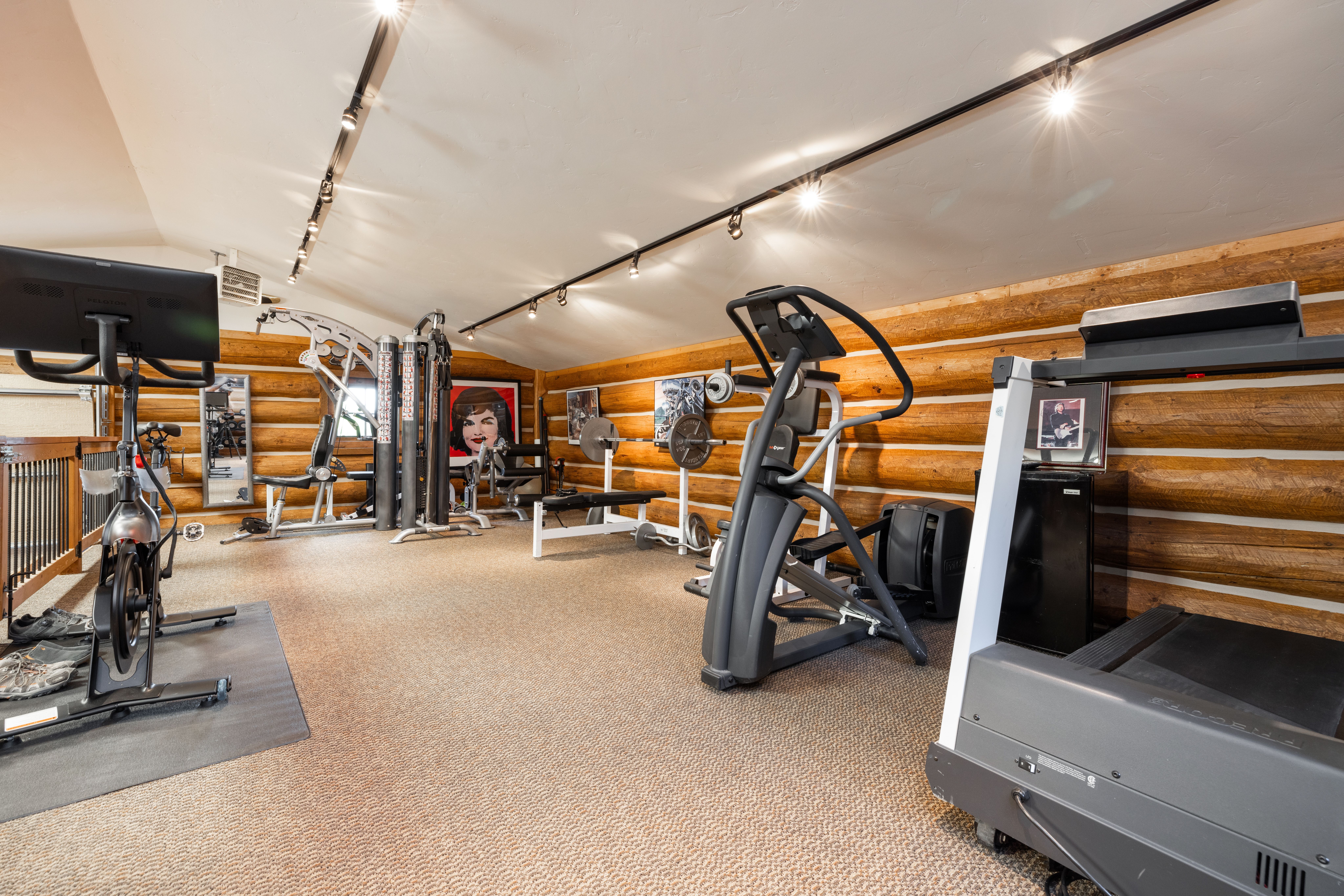 This spacious home gym features rustic log walls and a neutral carpeted floor, creating a warm and inviting workout environment. The room is well-equipped with a variety of fitness machines, including a Peloton bike, a multi-station weight system, a treadmill, and an elliptical trainer. Track lighting mounted on the slanted ceiling illuminates the space, which also includes framed artwork and a small refrigerator for convenience.