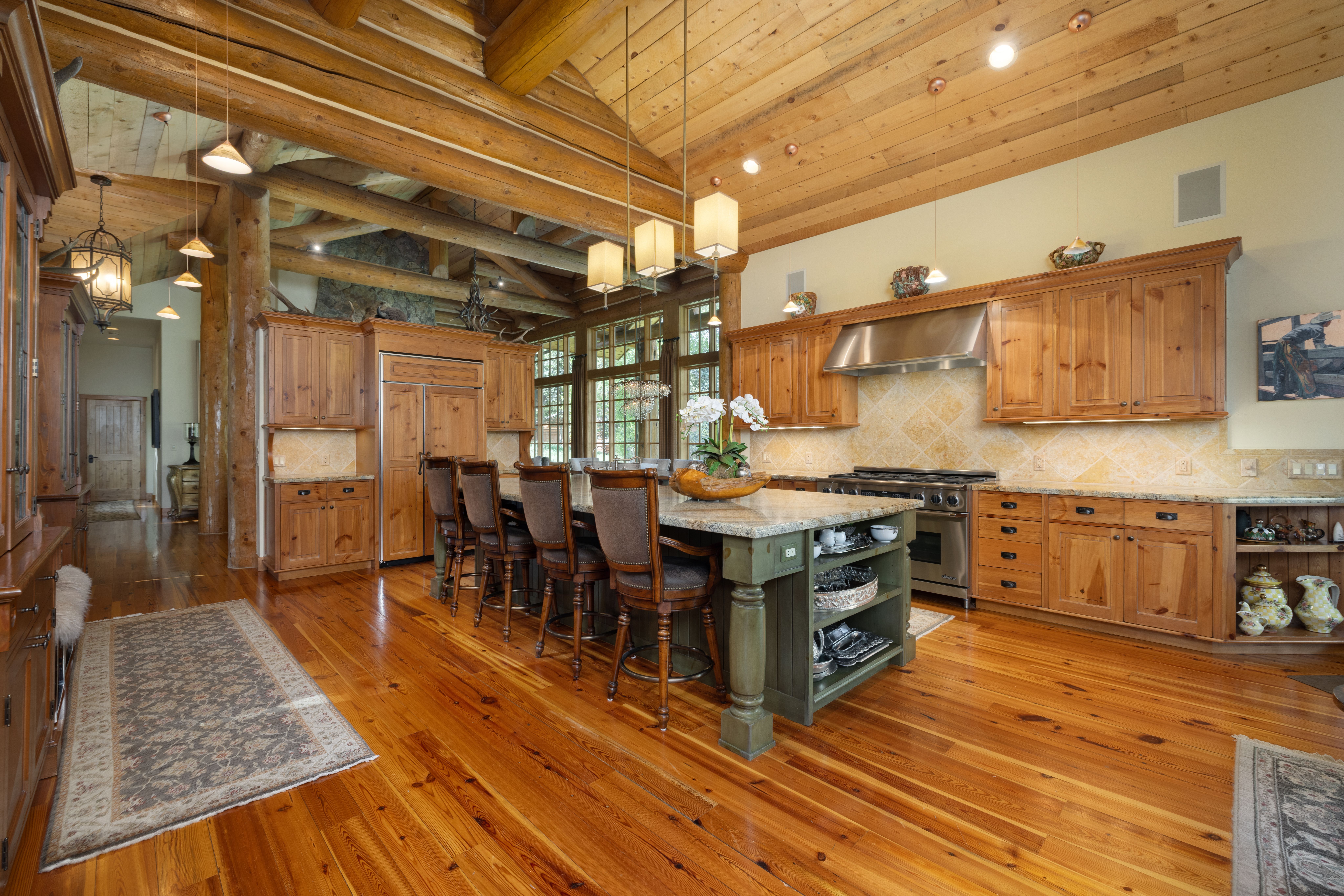 This rustic, high-end kitchen features warm wood cabinetry, exposed log beams, and a large central island with a green base and granite countertop. The space is illuminated by elegant pendant lighting and recessed lights, highlighting the rich hardwood floors and stone-tiled backsplash. The perspective is a wide-angle shot from the kitchen floor, showcasing the open-concept layout and the seamless blend of natural materials and modern appliances.