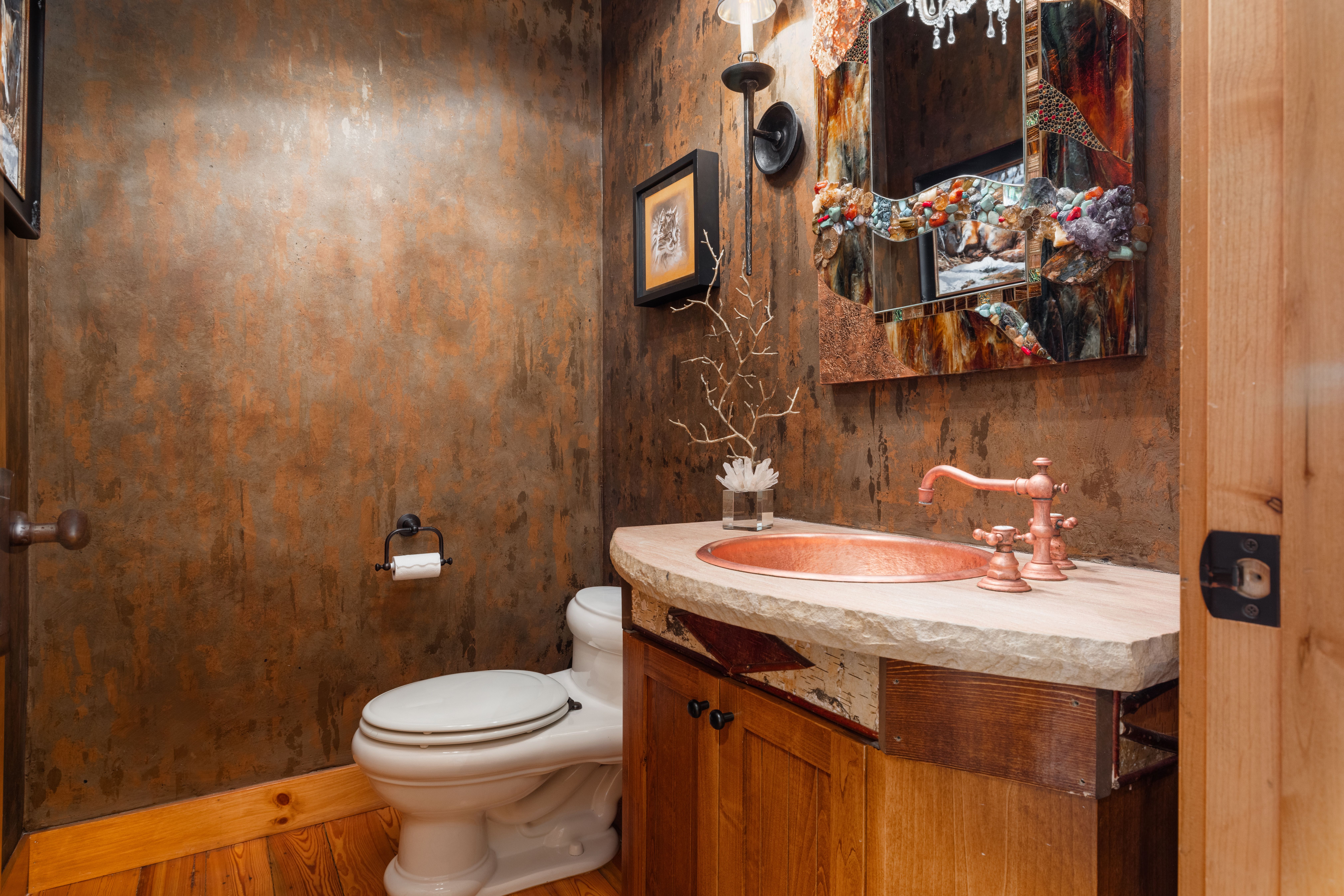 This unique powder room features a rustic, artisanal aesthetic with textured, copper-toned walls and a custom stone-topped vanity. The space is highlighted by a striking, crystal-encrusted mirror, a hammered copper sink, and a white porcelain toilet, all set against warm wood cabinetry and flooring. The overall impression is one of high-end, nature-inspired craftsmanship that creates a cozy and memorable atmosphere.
