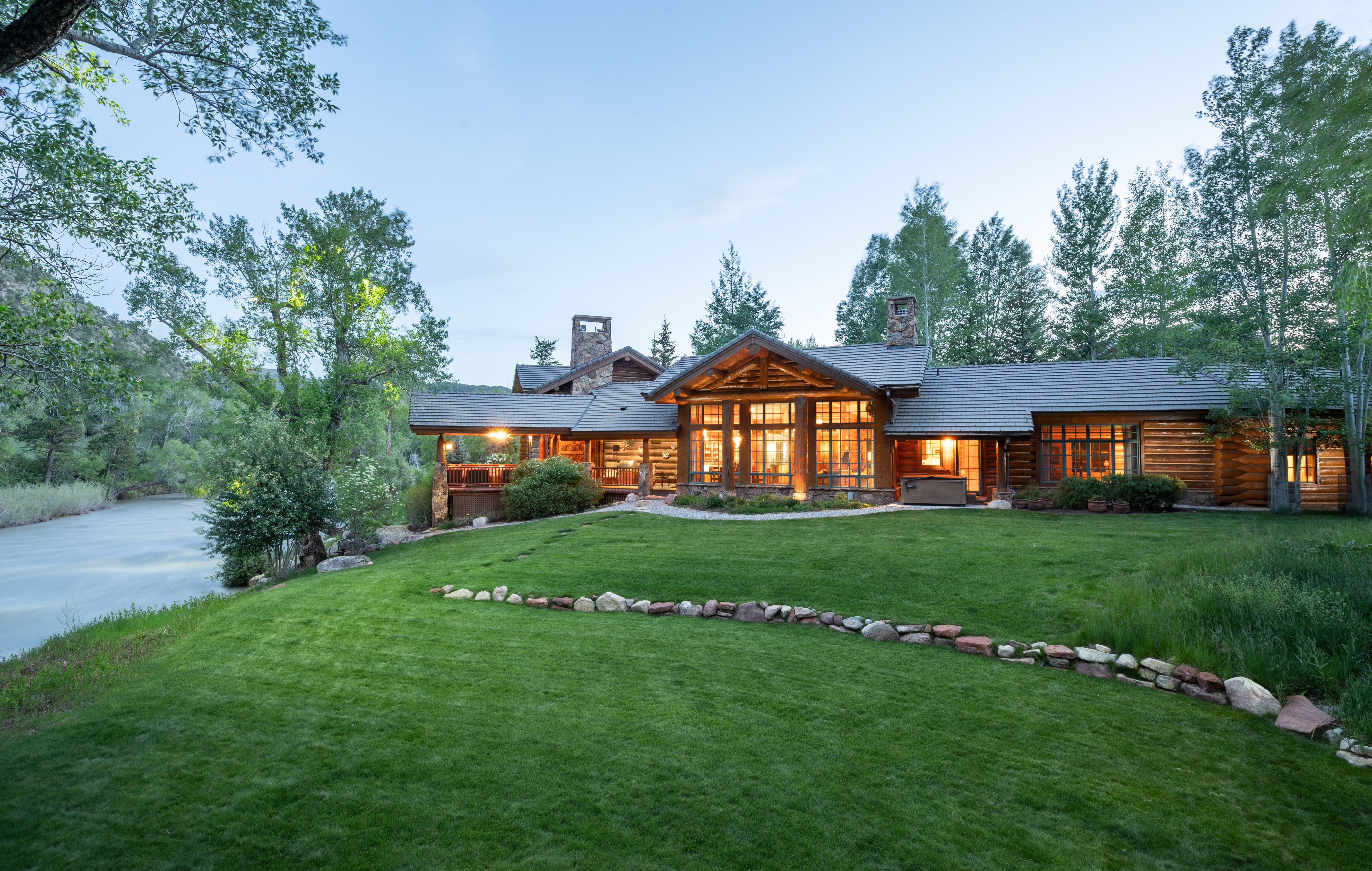 This stunning log cabin estate is captured from a low-angle perspective, showcasing its expansive rear lawn that slopes gently toward a flowing river. The home features rustic timber construction, large floor-to-ceiling windows that glow warmly in the twilight, and a spacious covered deck area. The serene natural setting, framed by lush trees and a stone-lined garden path, creates an inviting and luxurious mountain retreat atmosphere.