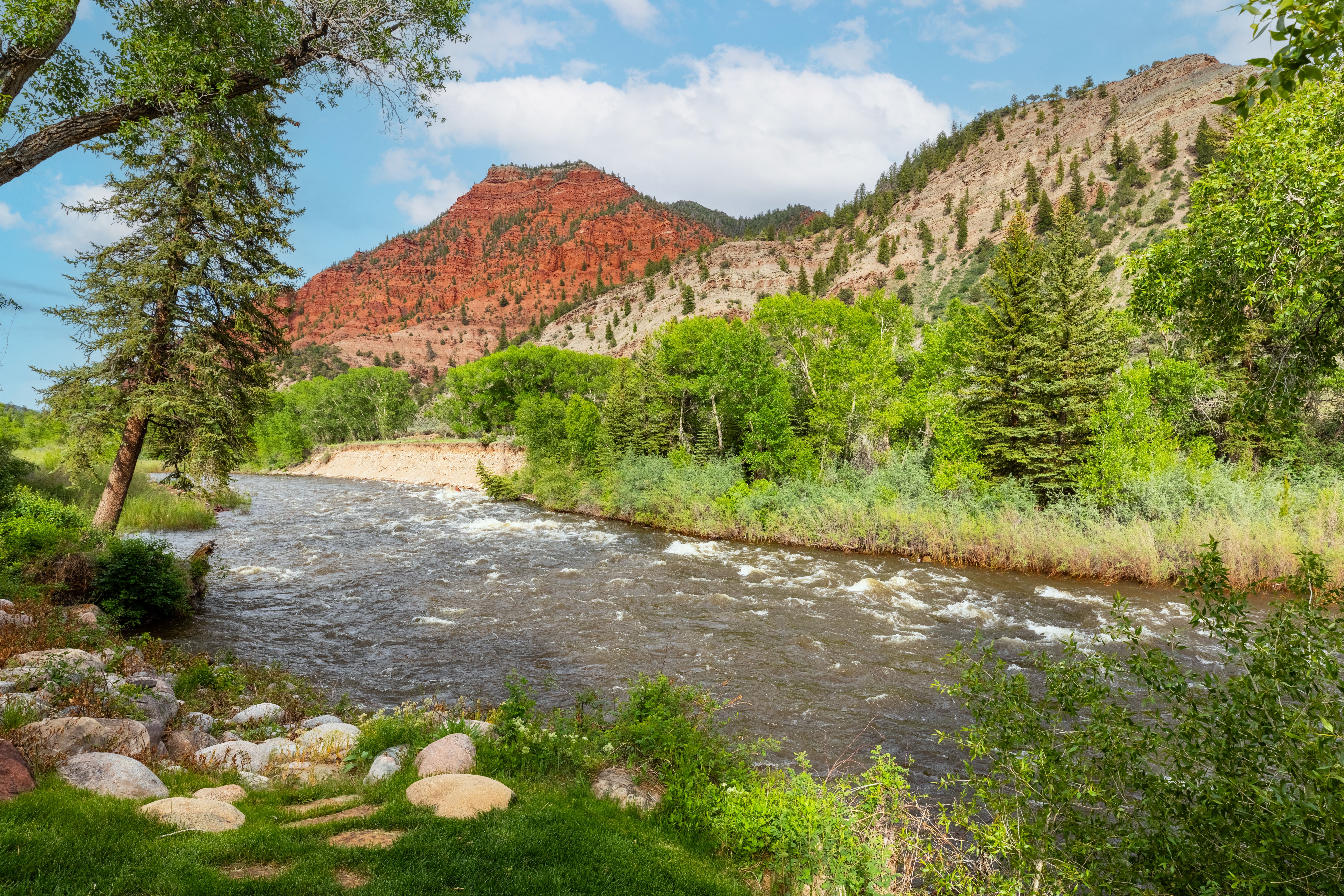 This scenic landscape captures a rushing river flowing through a lush, green valley framed by towering, rugged red rock mountains. The foreground features a grassy bank with scattered river stones, while the mid-ground is filled with vibrant deciduous trees and evergreens. The perspective is a wide-angle, eye-level shot that emphasizes the natural beauty and serene atmosphere of the property's surroundings.