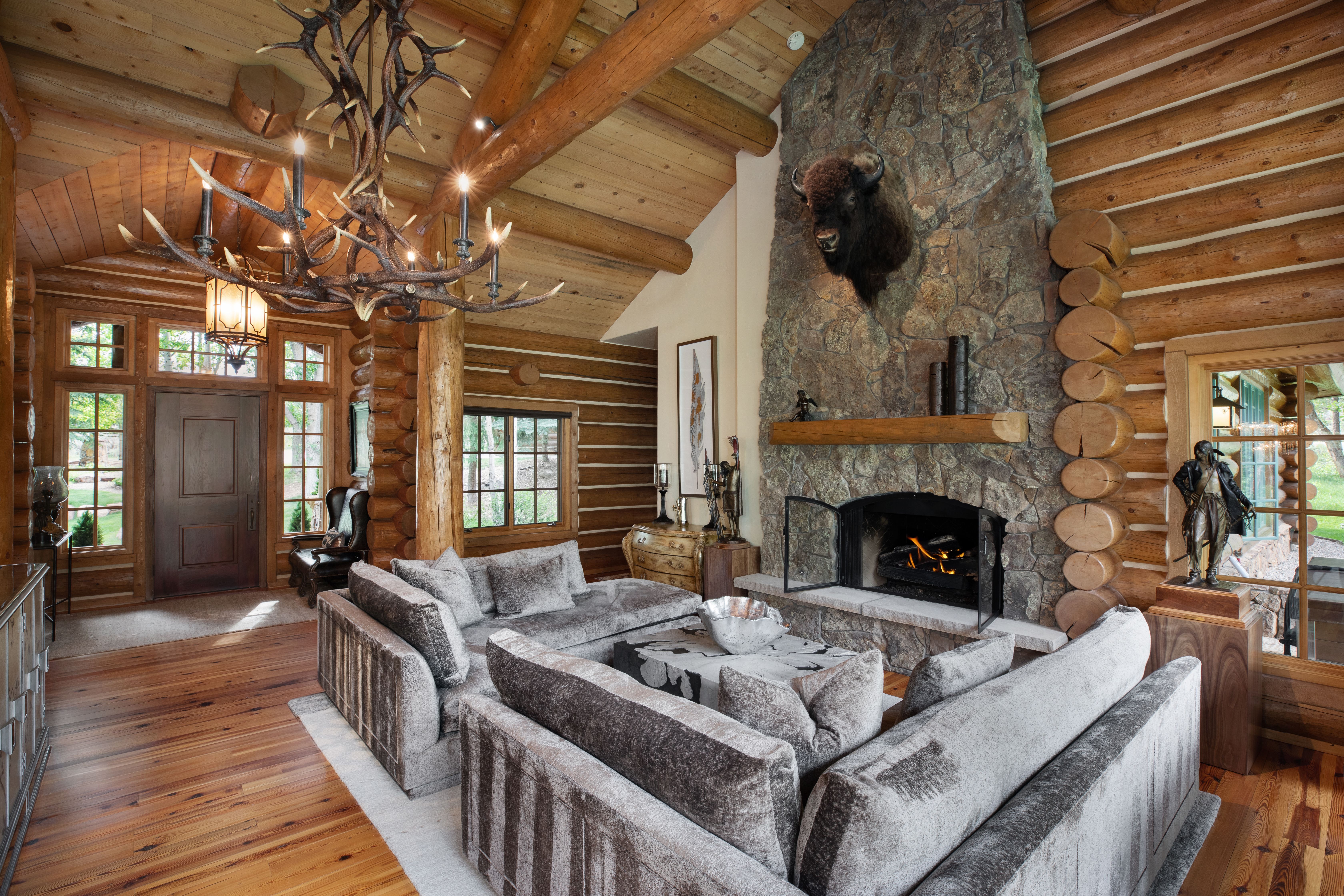 This rustic living room features a grand stone fireplace as its focal point, adorned with a bison taxidermy mount and a heavy timber mantel. The space is defined by exposed log walls, vaulted ceilings with massive timber beams, and a striking antler chandelier that adds a dramatic, lodge-style aesthetic. Plush, textured grey sofas are arranged around a contemporary coffee table, creating a cozy and inviting atmosphere perfect for a mountain retreat.