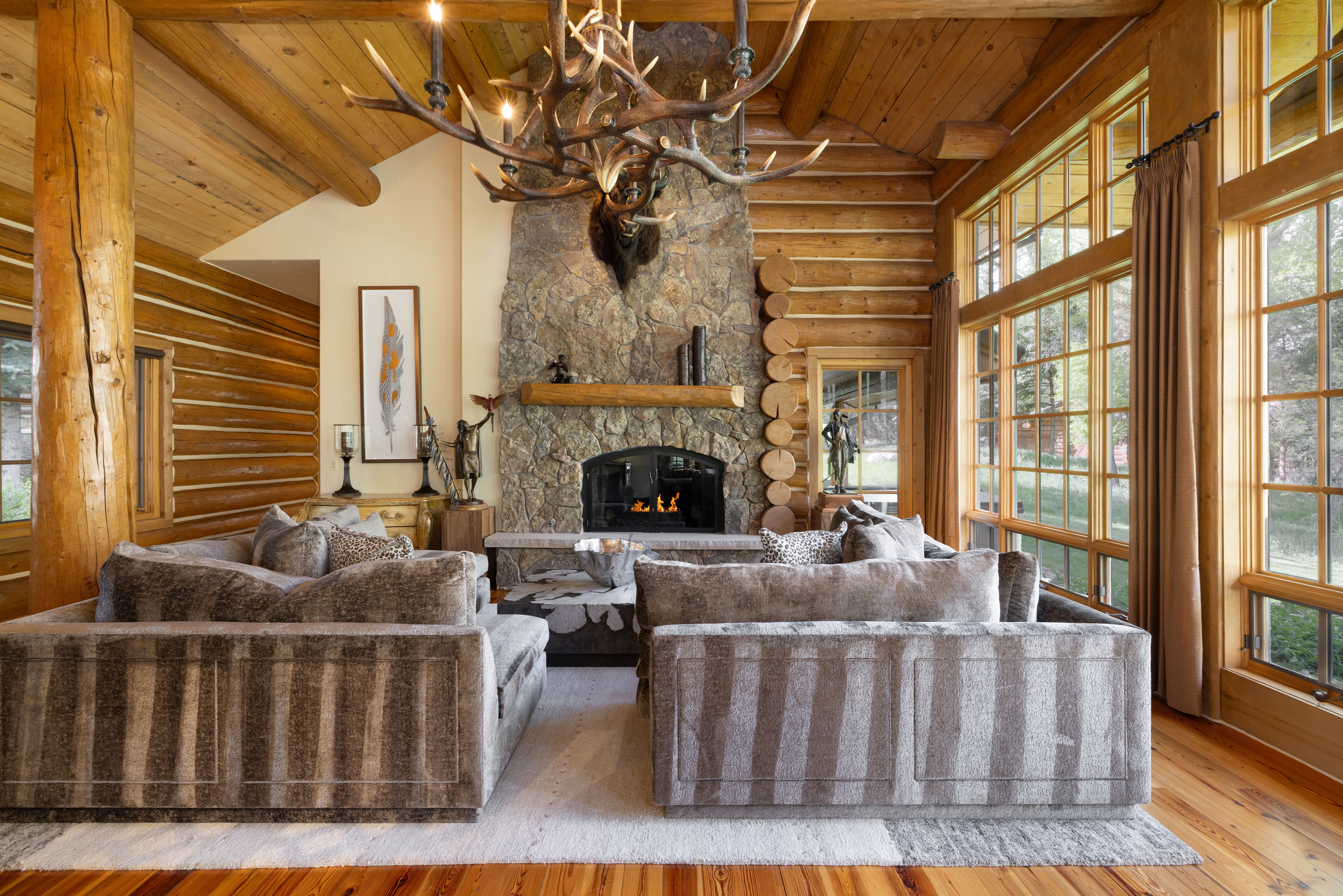 This rustic living room features a grand stone fireplace as its centerpiece, adorned with a large antler chandelier and exposed log walls. The space is furnished with plush, textured sofas arranged around a central coffee table, creating a cozy and inviting atmosphere. Large windows allow natural light to flood the room, highlighting the warm wood tones and high-end mountain lodge aesthetic.