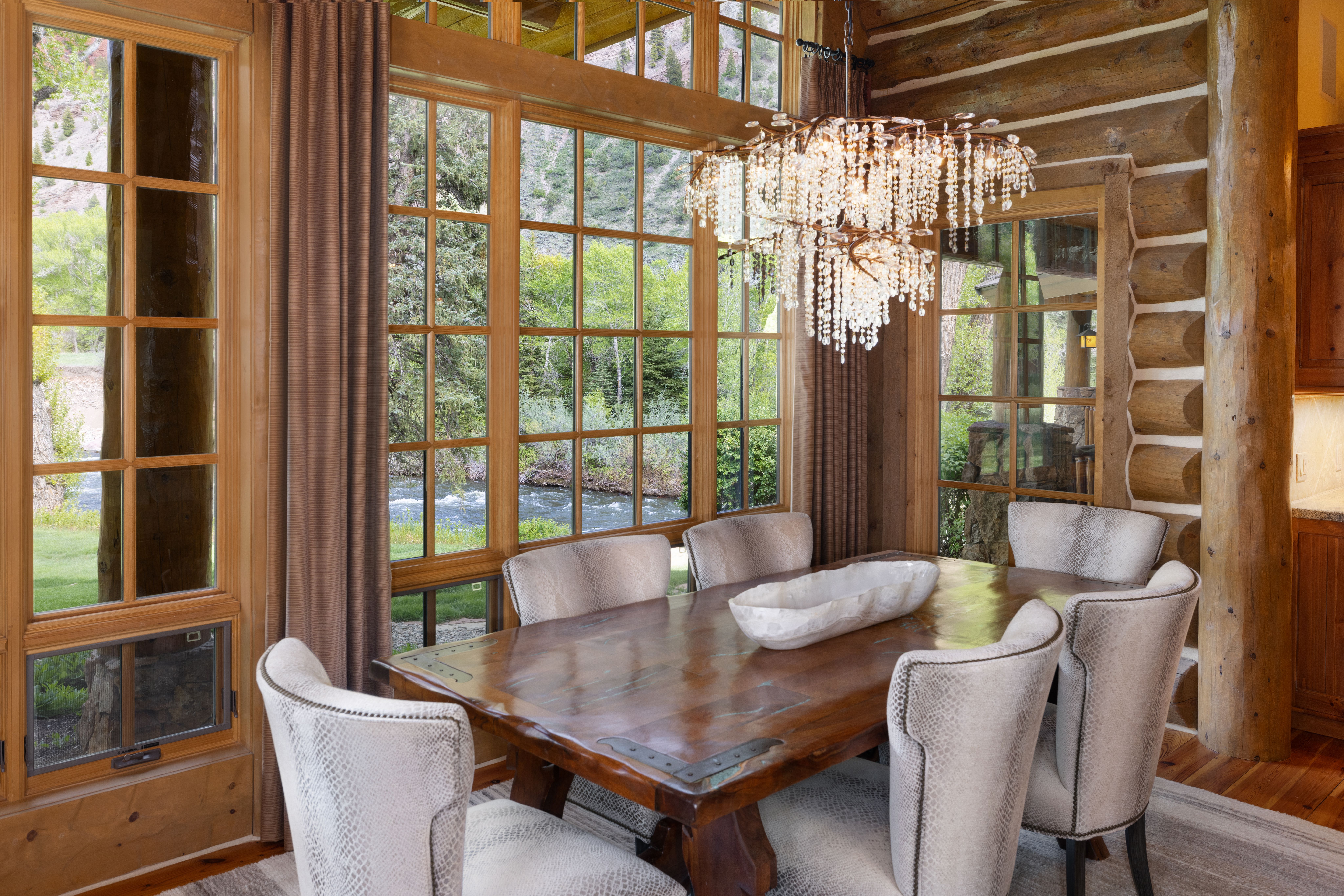 This elegant dining room features a rustic log cabin aesthetic with exposed timber walls and a large wooden table accented by metal corner plates. A stunning crystal chandelier hangs above a central stone bowl centerpiece, while floor-to-ceiling windows provide a picturesque view of a flowing river and lush forest. The space is furnished with sophisticated, textured upholstered chairs, creating a warm and luxurious atmosphere perfect for scenic dining.