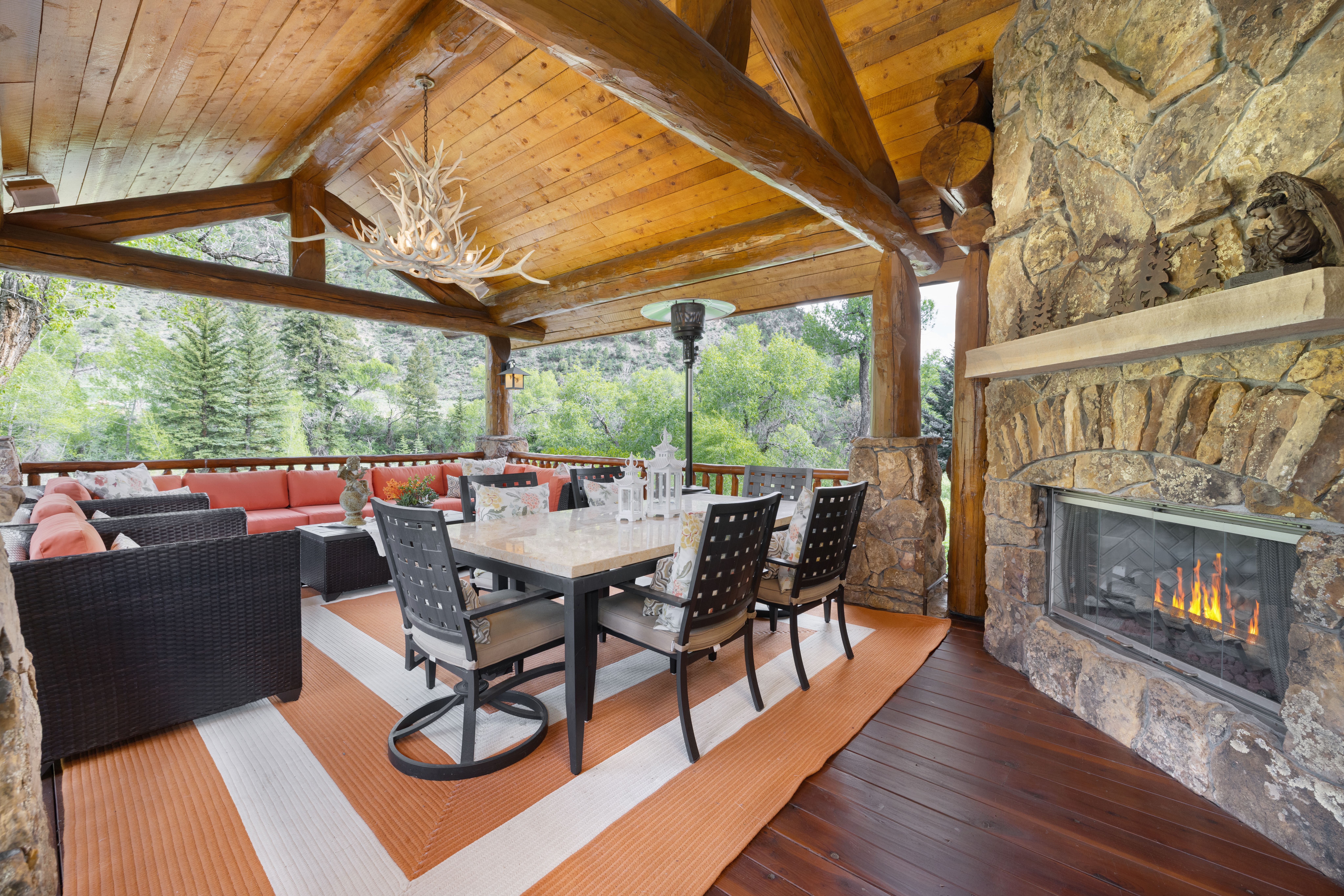 This expansive covered outdoor deck features a rustic log-cabin aesthetic with exposed timber beams and a massive stone fireplace. The space is furnished with a comfortable sectional sofa and a formal dining set, all set atop a vibrant orange and white striped rug. The open-air design provides a seamless transition to the surrounding forest, creating an inviting and luxurious mountain retreat atmosphere.