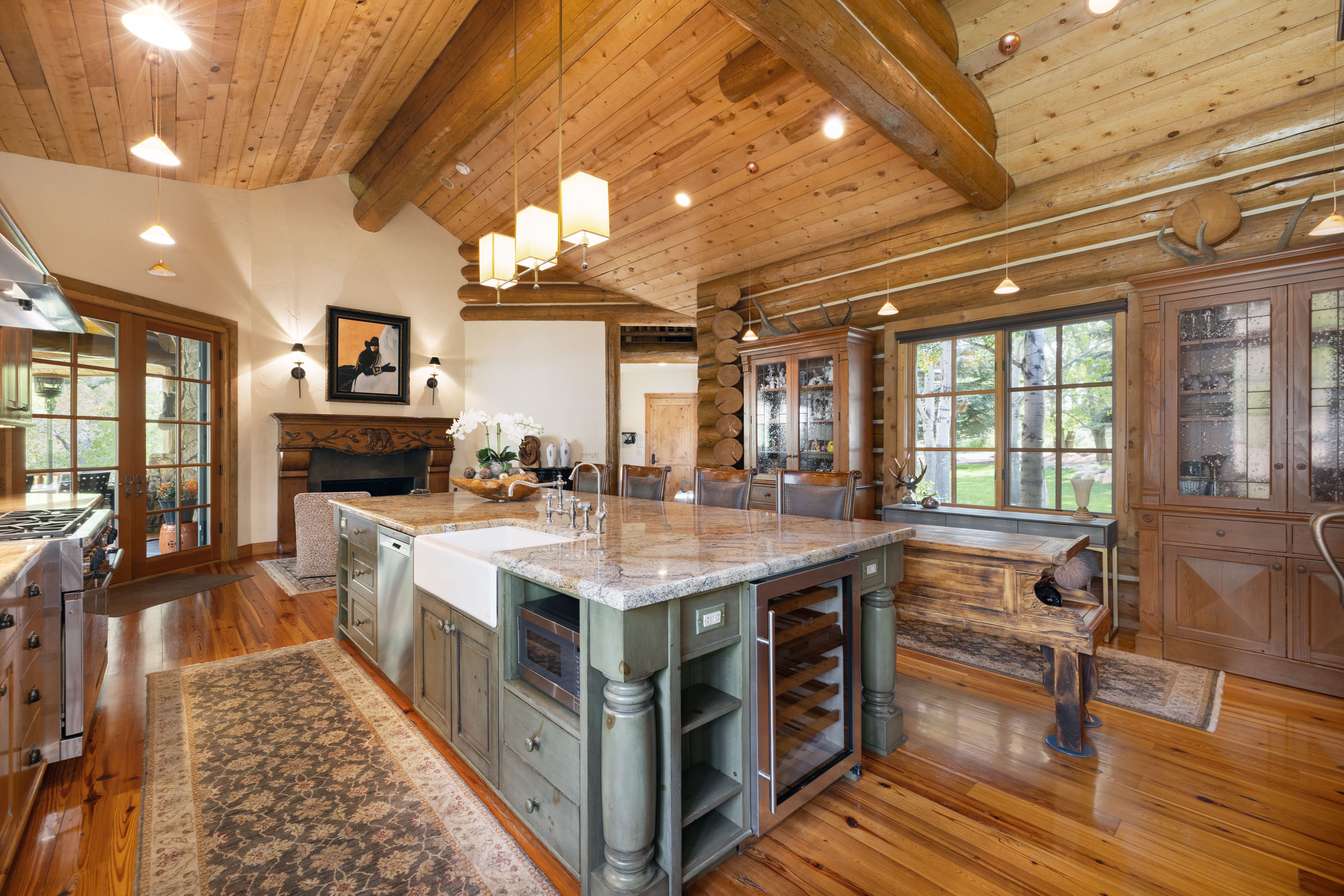 This rustic, high-end kitchen features a large central island with a farmhouse sink, granite countertops, and a built-in wine cooler, all set against a backdrop of warm log walls and exposed timber beams. The space is anchored by a stone fireplace and elegant cabinetry, creating a cozy yet sophisticated atmosphere. The perspective is a wide-angle shot from the kitchen floor, showcasing the seamless blend of natural wood textures and modern luxury amenities.