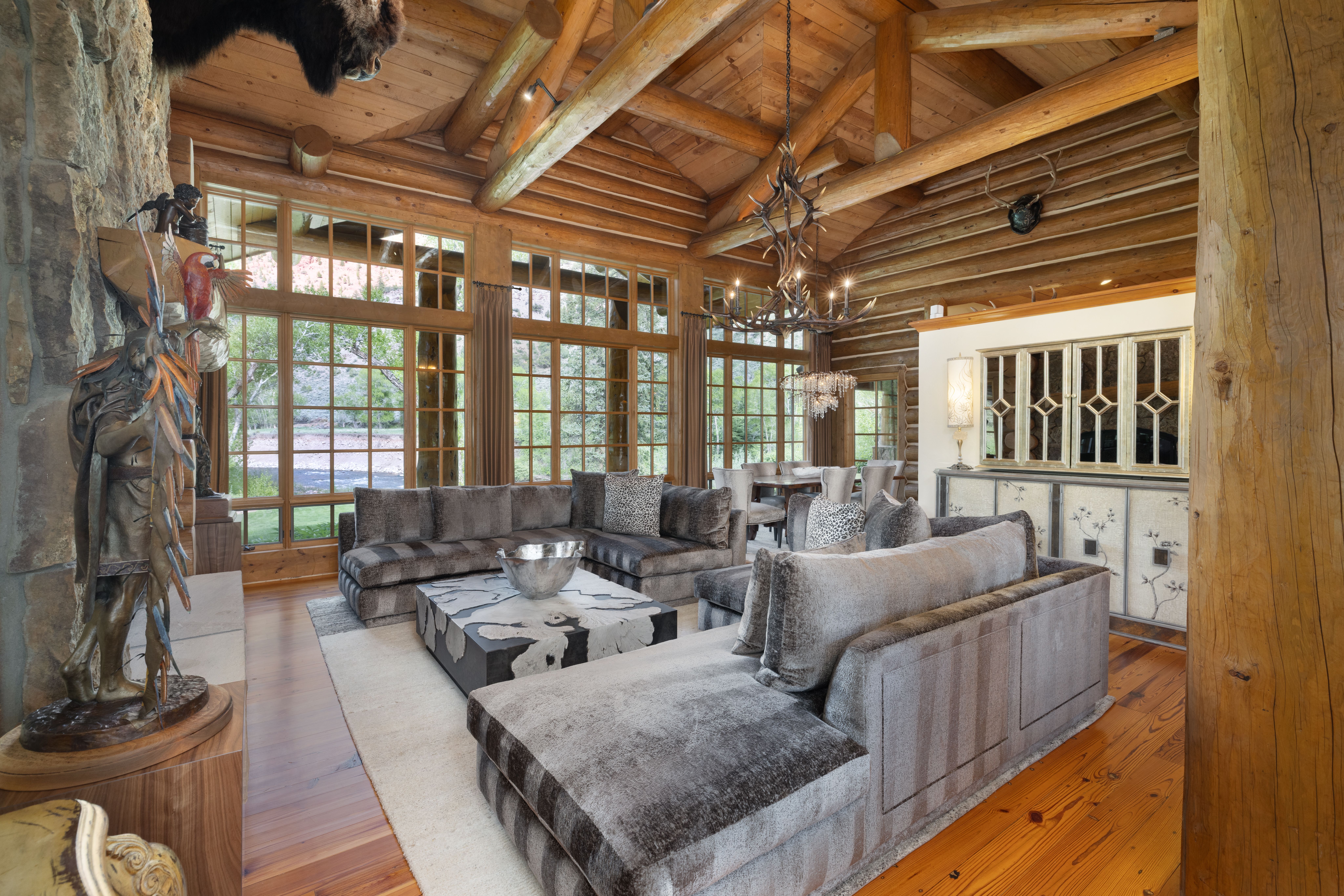 This expansive, rustic living room features high vaulted ceilings with exposed log beams and a massive stone fireplace, creating a warm and luxurious mountain lodge atmosphere. The space is anchored by a large, plush sectional sofa and a unique, patterned coffee table, all set against a backdrop of floor-to-ceiling windows that offer scenic views of the outdoors. An antler chandelier serves as a striking focal point, enhancing the sophisticated, cabin-inspired aesthetic of the home.