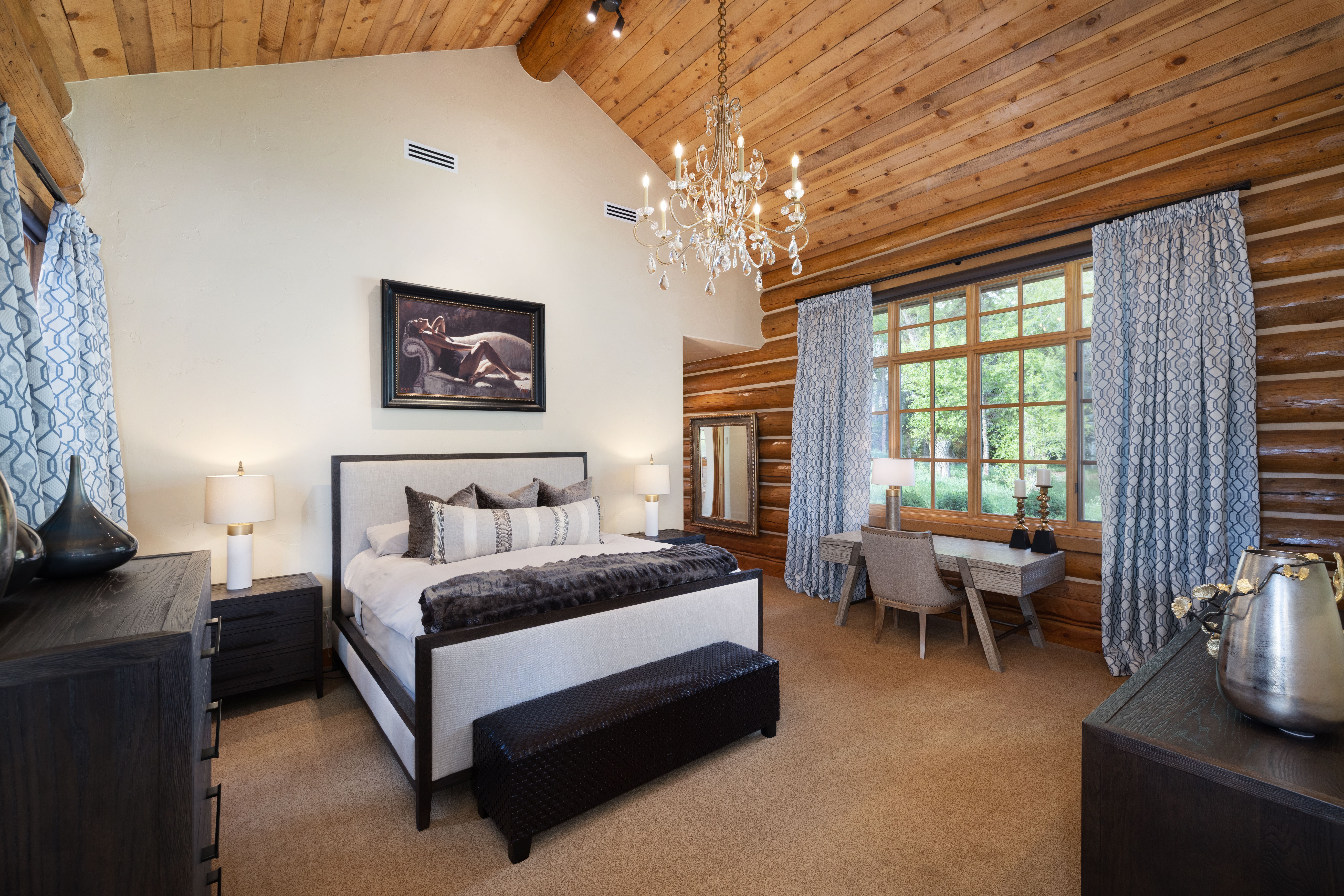 This luxurious primary bedroom features a rustic log-cabin aesthetic with exposed timber ceilings and walls, balanced by a sophisticated, modern interior design. The room is anchored by a large upholstered bed with a dark wood frame, complemented by a matching bench, nightstands, and a dedicated workspace near the window. A grand crystal chandelier serves as a focal point, while the neutral color palette and plush carpeting create a warm, inviting, and high-end atmosphere.