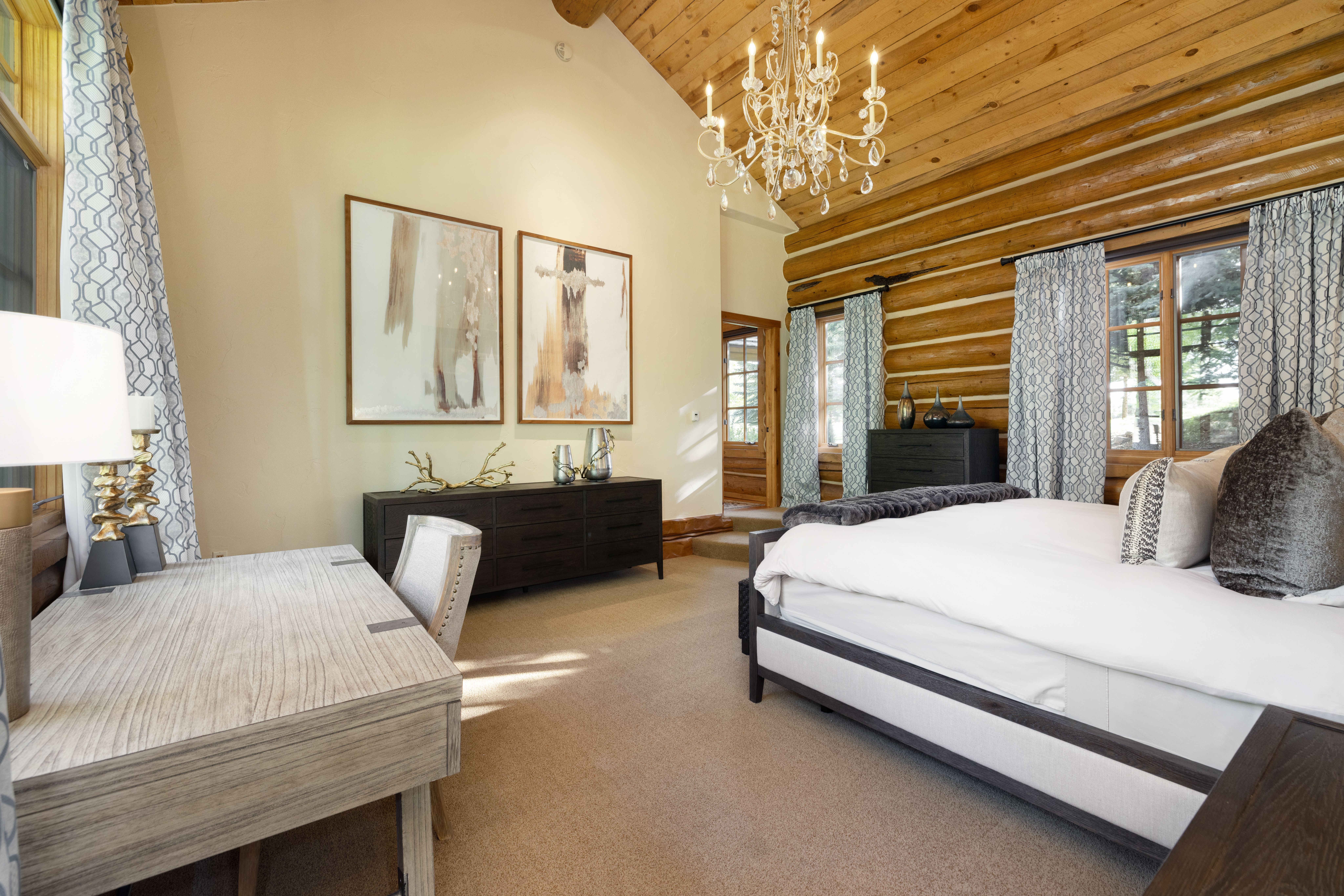 This spacious primary bedroom features a rustic log cabin aesthetic with exposed timber walls and a vaulted wood-paneled ceiling. The room is elegantly furnished with a large bed, a modern desk, and a dark wood dresser, all illuminated by a sophisticated crystal chandelier. Large windows draped in patterned curtains allow natural light to fill the space, creating a warm and inviting atmosphere.