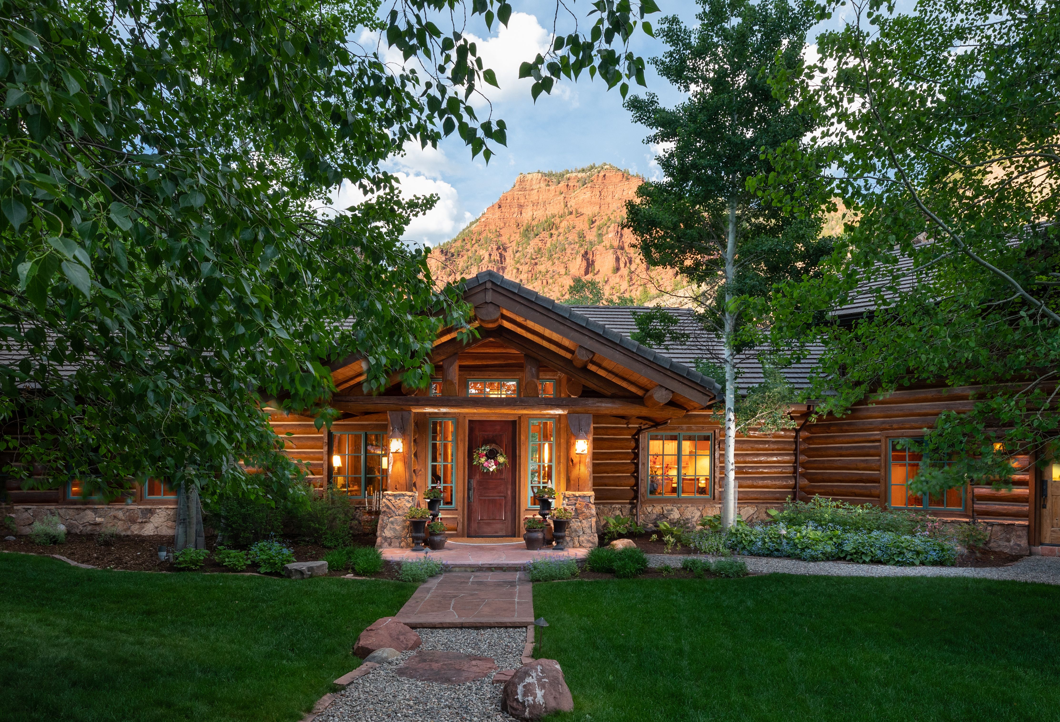 This stunning log cabin home features a rustic architectural style with natural stone accents and warm, inviting exterior lighting. The front entryway is framed by lush green trees and a well-manicured lawn, with a dramatic red rock mountain peak rising in the background. The perspective is a centered, eye-level shot that emphasizes the home's harmonious integration with its scenic, mountainous environment.