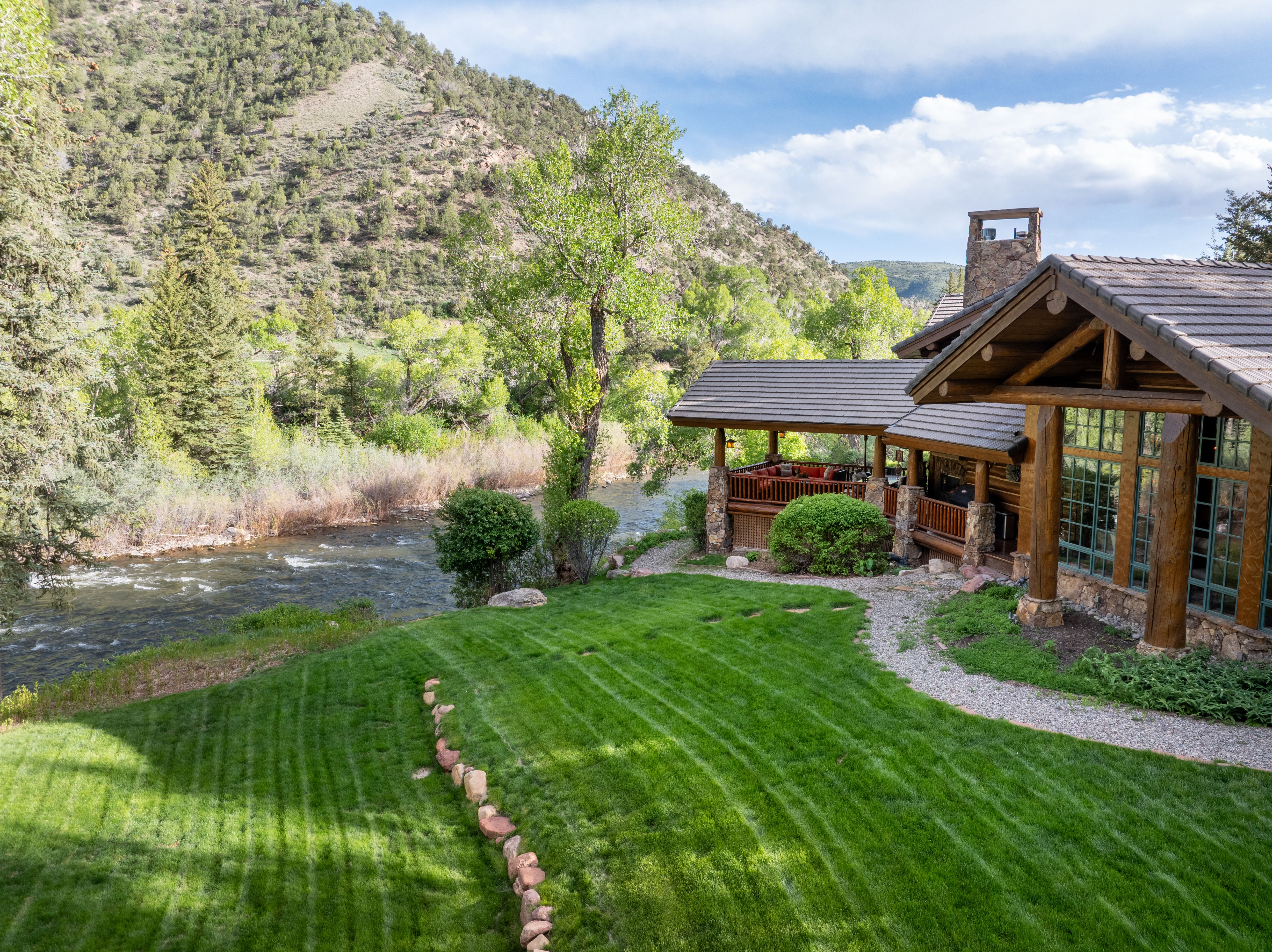 This elevated perspective captures a stunning log-style mountain home nestled alongside a rushing river. The property features a lush, manicured lawn with a stone-lined path leading toward a spacious covered deck, all set against a backdrop of towering pine trees and rugged hills. The scene evokes a serene, high-end retreat atmosphere, perfectly blending rustic architectural charm with the natural beauty of the landscape.