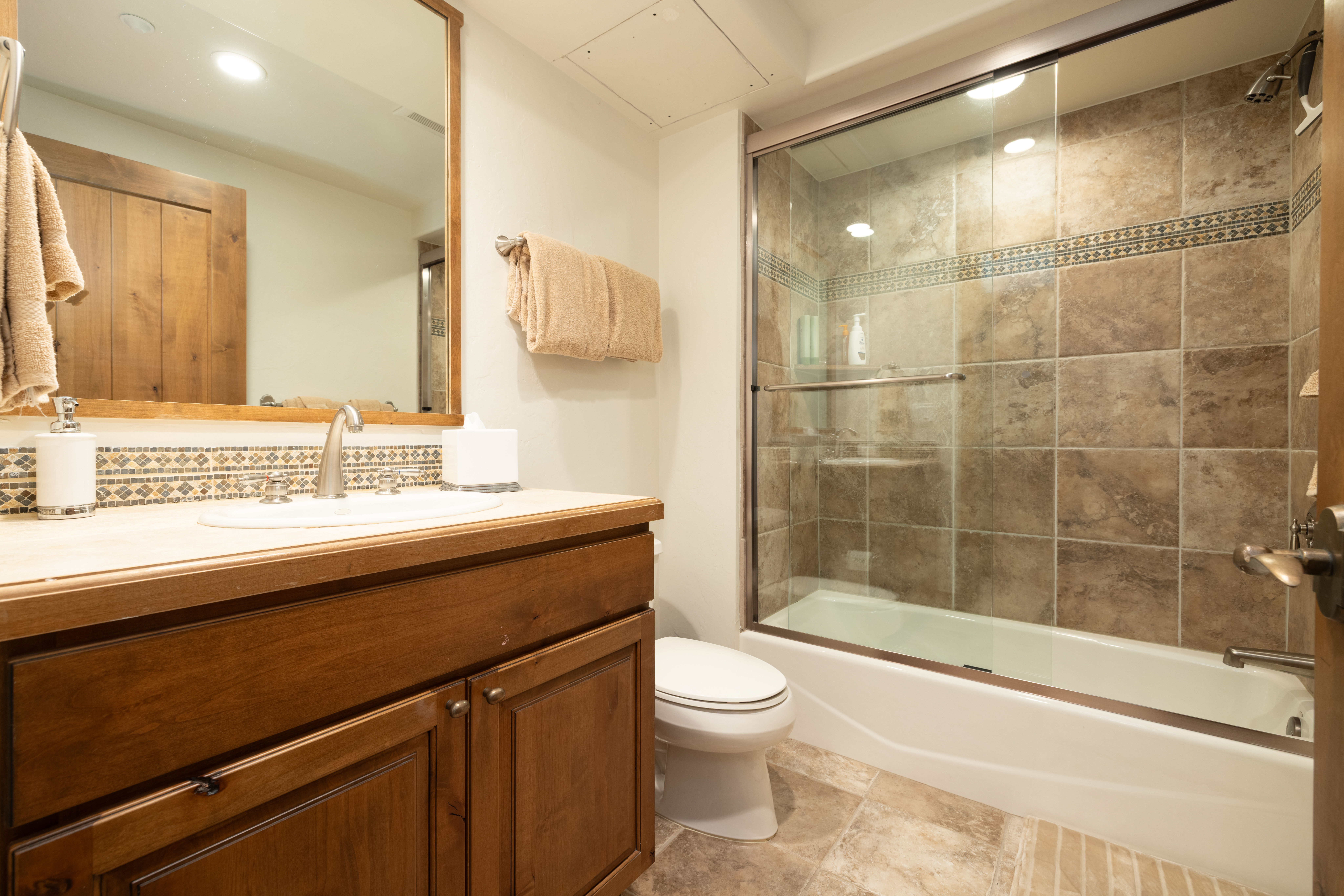 This bathroom features a rustic aesthetic with a warm-toned wood vanity, a stone-topped counter, and a matching framed mirror. The shower-tub combination is enclosed by sliding glass doors and finished with neutral-toned square tiles and a decorative mosaic accent band. The space is completed by a white toilet and light-colored floor tiles, creating a cohesive and functional design.