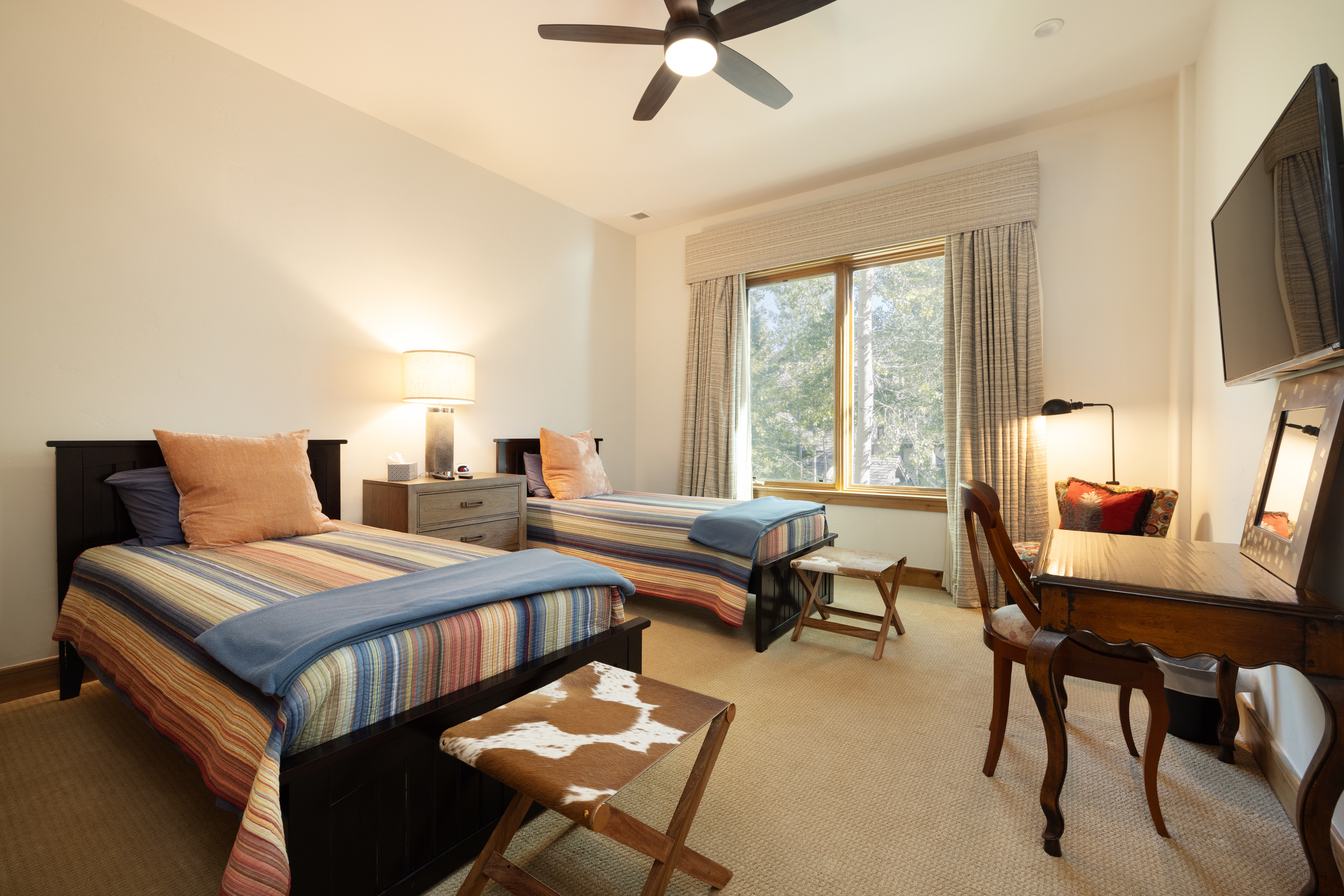 This guest bedroom features two twin beds with striped bedding and solid blue throws, centered around a shared nightstand with a table lamp. A wooden desk with a chair and a wall-mounted television are positioned to the right, while a cowhide-patterned stool sits at the foot of the bed in the foreground. The room is illuminated by a ceiling fan light and natural light streaming through a large window framed by patterned curtains.