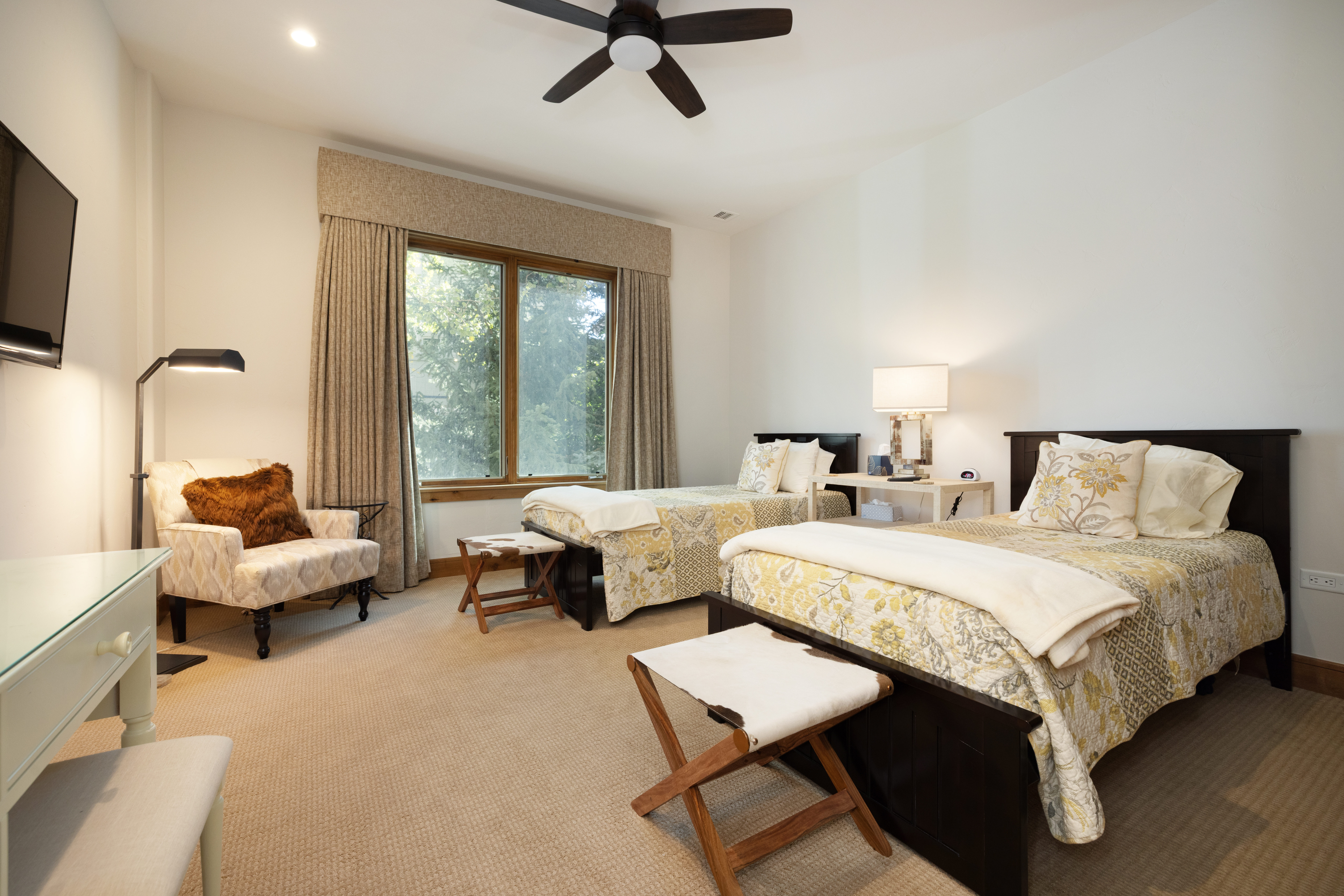 This well-appointed guest bedroom features two twin beds with dark wood frames and patterned yellow and white bedding. The room includes a cozy armchair with a faux-fur pillow, a small desk area, and two wooden stools with hide-patterned seats placed at the foot of each bed. Large windows with neutral drapery allow natural light to fill the space, which is finished with wall-to-wall carpeting and a ceiling fan.