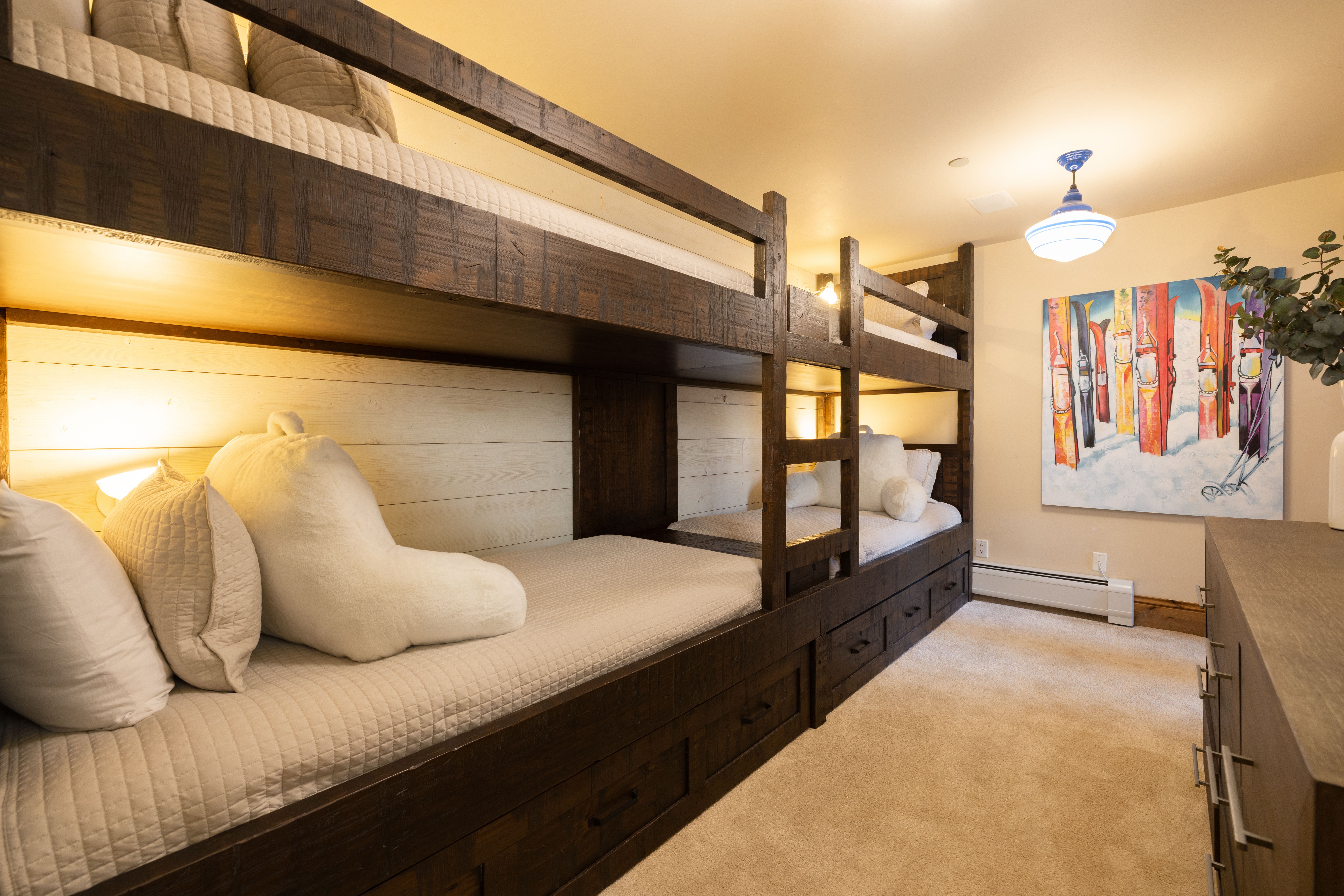 This cozy, rustic-style bunk room features custom-built dark wood bunk beds with integrated storage drawers, perfect for maximizing space. The room is accented with warm, ambient lighting, plush white bedding, and a vibrant piece of ski-themed wall art that adds a playful touch. The perspective is a wide-angle shot from the foot of the beds, emphasizing the comfortable and inviting atmosphere of this mountain-inspired retreat.
