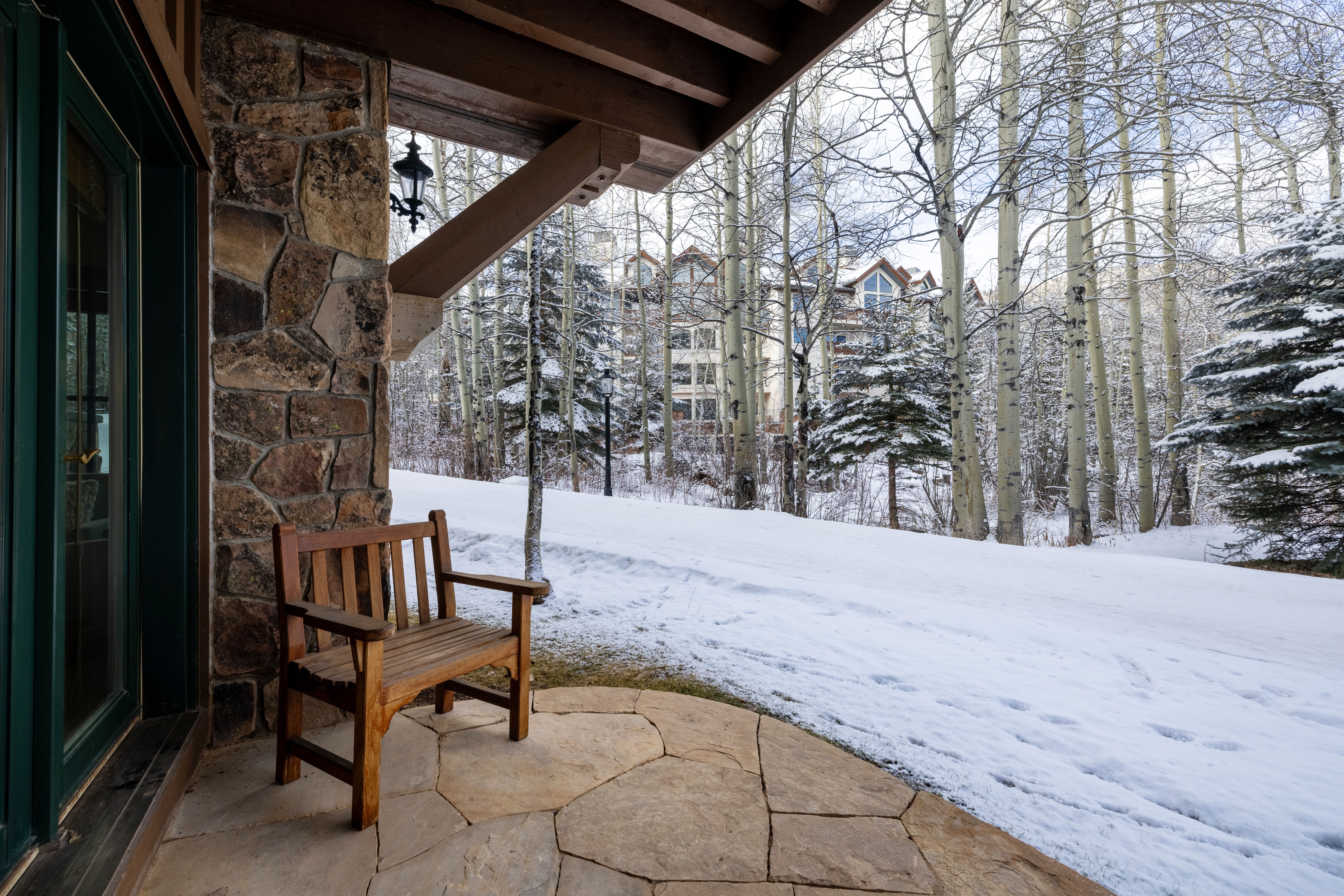 This inviting stone patio features a classic wooden bench, offering a serene spot to enjoy the snowy, wooded landscape. The architectural details include rustic stone masonry and a sturdy timber-framed overhang, creating a cozy, mountain-lodge aesthetic. The perspective looks out from the covered porch toward a snow-covered path and a multi-story building nestled among tall, bare aspen trees.