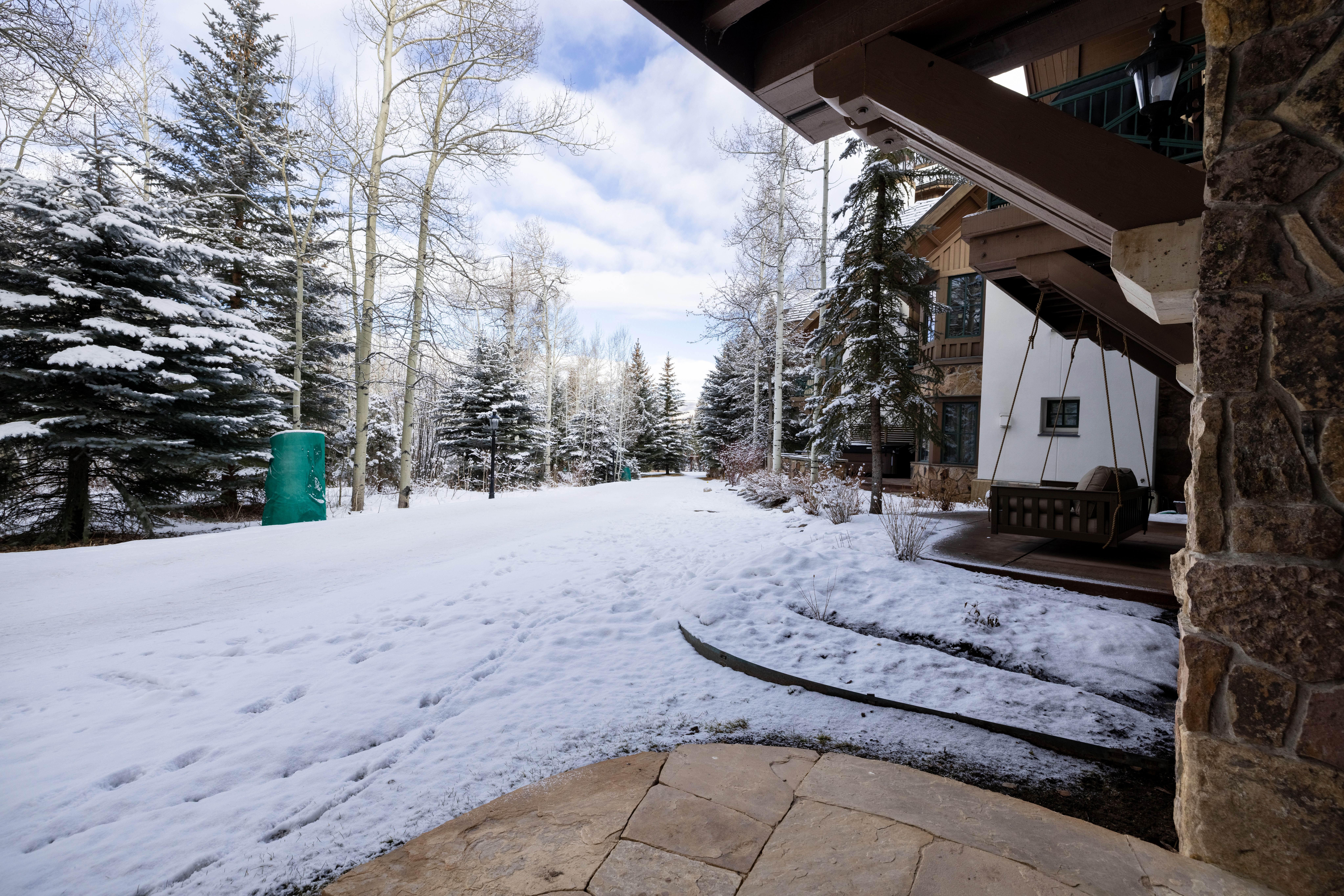 This image captures a serene, snow-covered patio area featuring a rustic wooden porch swing suspended under a covered entryway. The scene is framed by a stone pillar in the foreground and opens up to a winter landscape filled with snow-dusted pine and aspen trees. The perspective provides a cozy, sheltered view looking out onto a quiet, wintry path, emphasizing the peaceful and secluded atmosphere of the property.