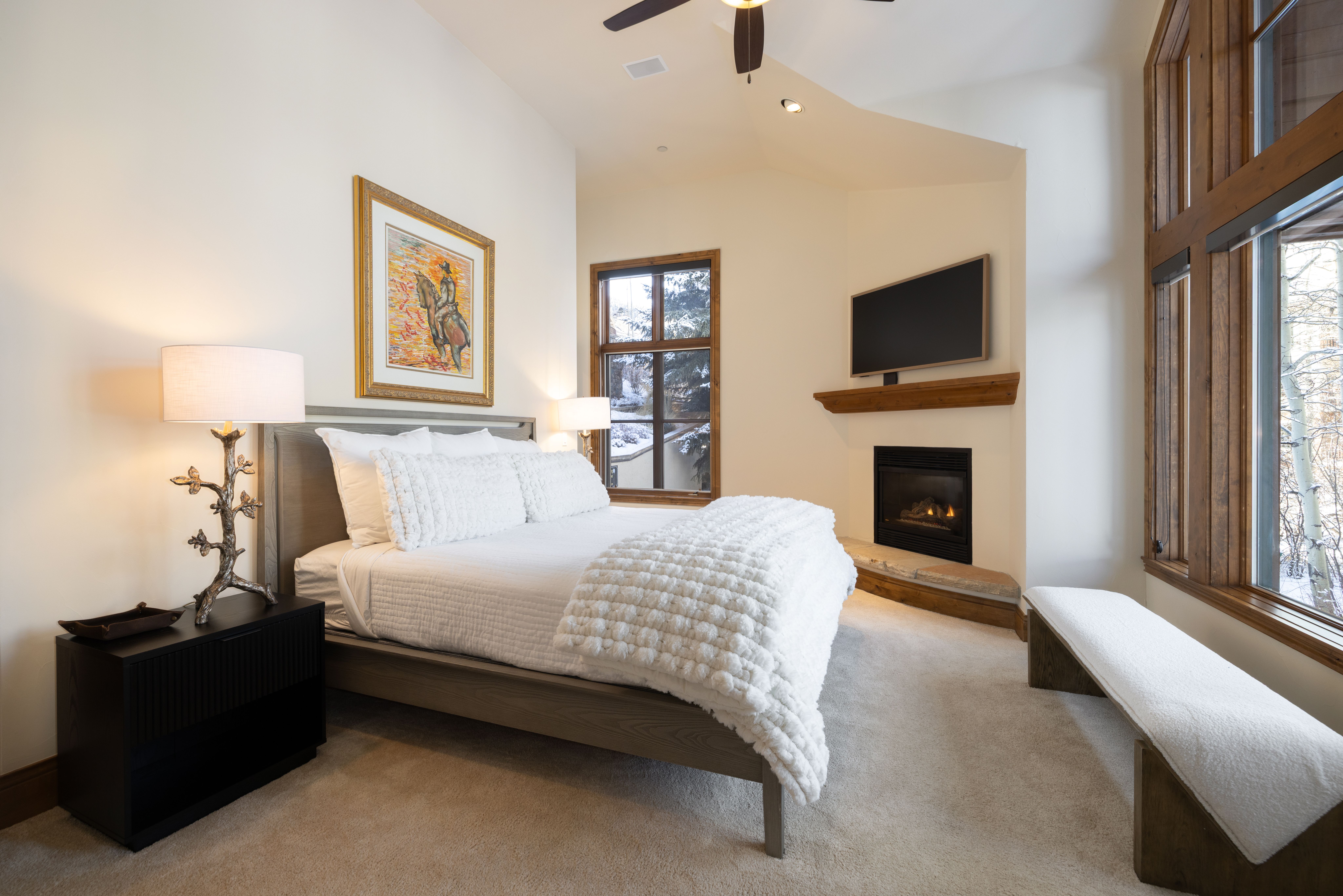 This cozy primary bedroom features a neutral color palette, a plush white textured bedspread, and a rustic wooden bed frame. A fireplace with a stone hearth and a mounted television creates a warm focal point, while large windows offer a serene view of a snowy landscape. The room is accented by a unique tree-branch style lamp and a framed western-themed artwork, providing a sophisticated mountain-retreat aesthetic.