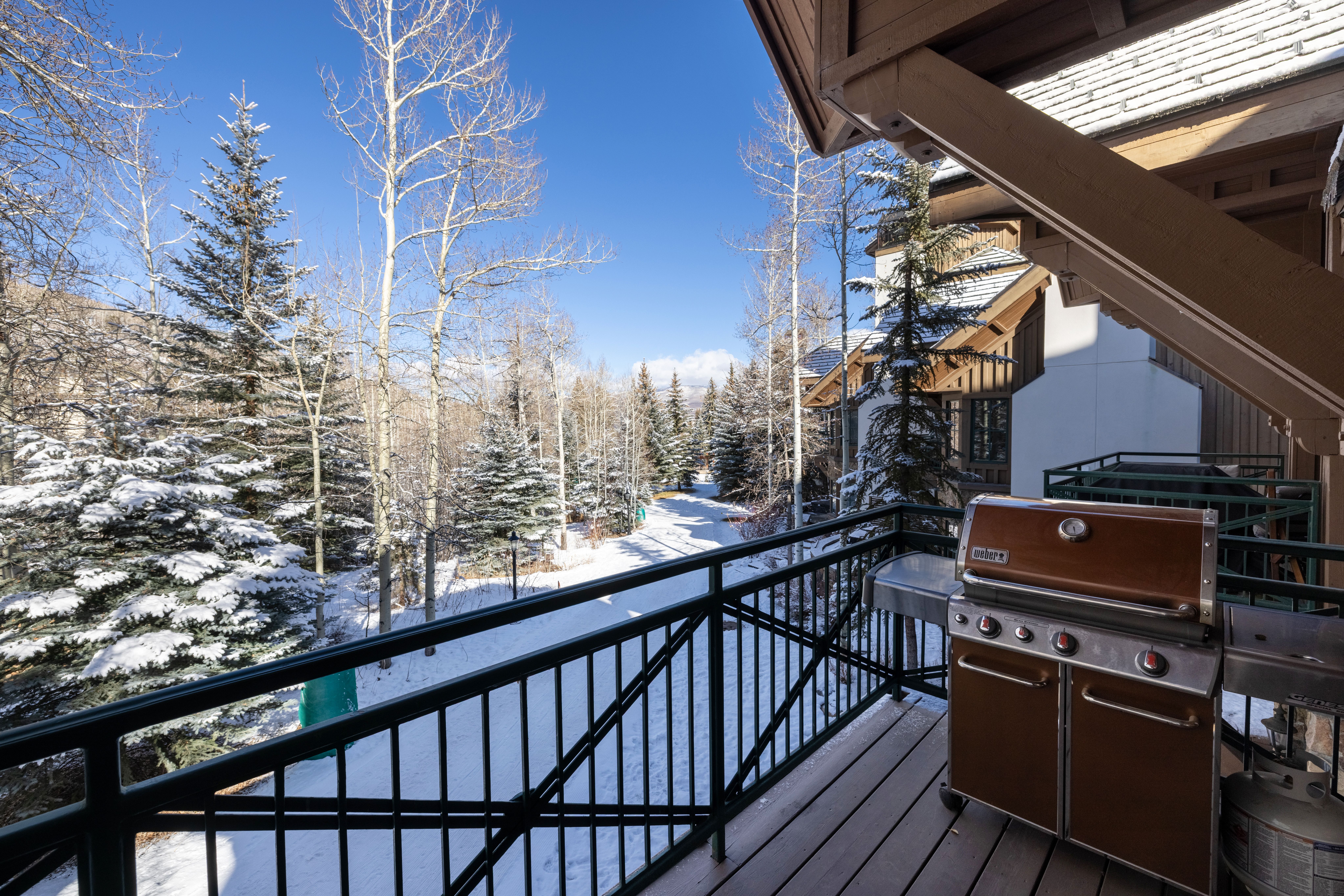 This elevated balcony offers a serene, wintery view of a snow-covered landscape surrounded by evergreen and aspen trees. The deck features a high-quality Weber grill, providing a perfect space for outdoor cooking while enjoying the crisp mountain atmosphere. The perspective captures the inviting transition from the cozy wooden deck to the expansive, tranquil forest beyond.