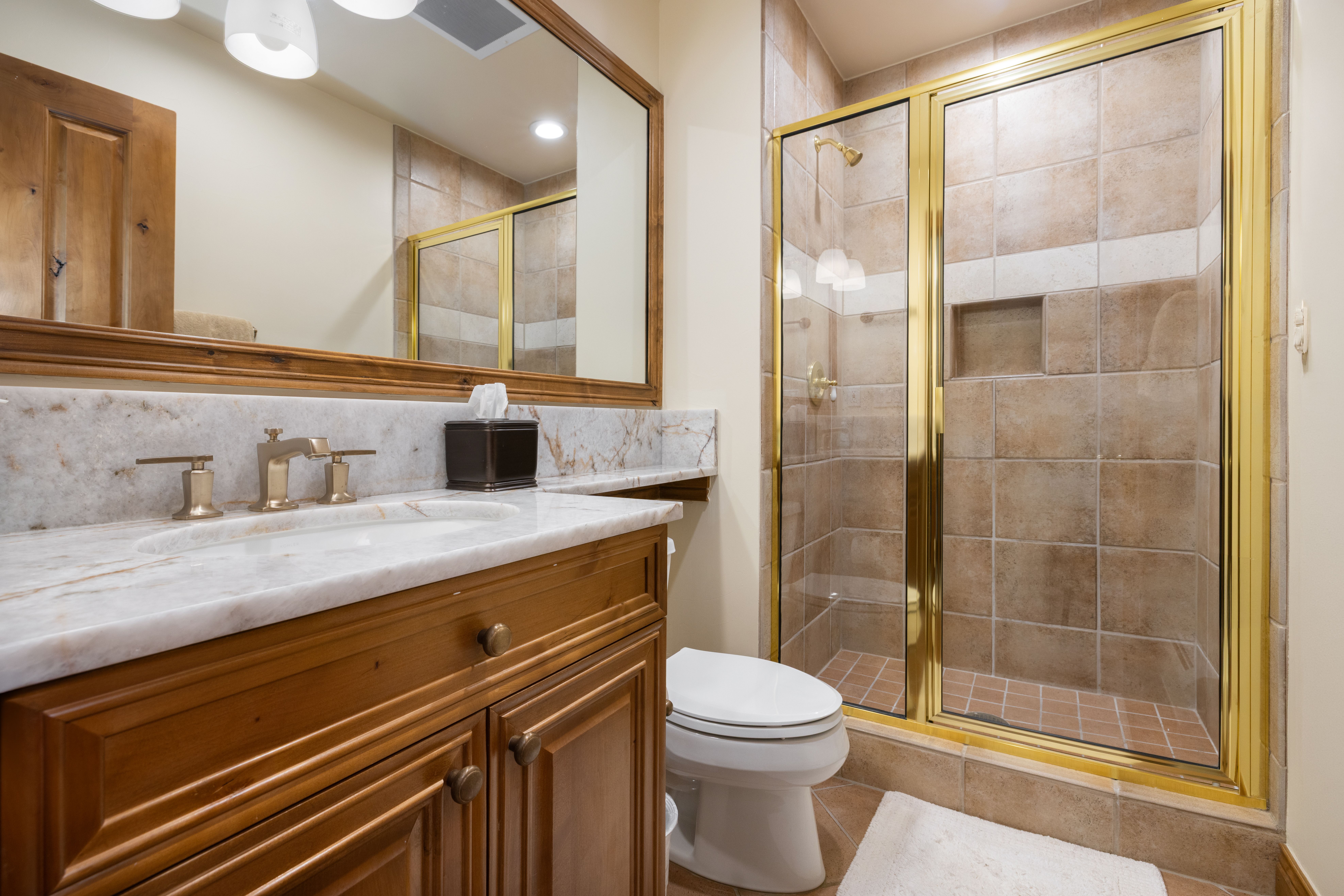 This compact bathroom features a warm, traditional aesthetic with a wooden vanity, marble countertop, and gold-toned fixtures. A glass-enclosed shower with tan tiled walls and a built-in niche sits adjacent to a white toilet, creating a functional and cohesive space. The perspective is a straight-on shot that captures the vanity, mirror, and shower area, highlighting the classic design elements.